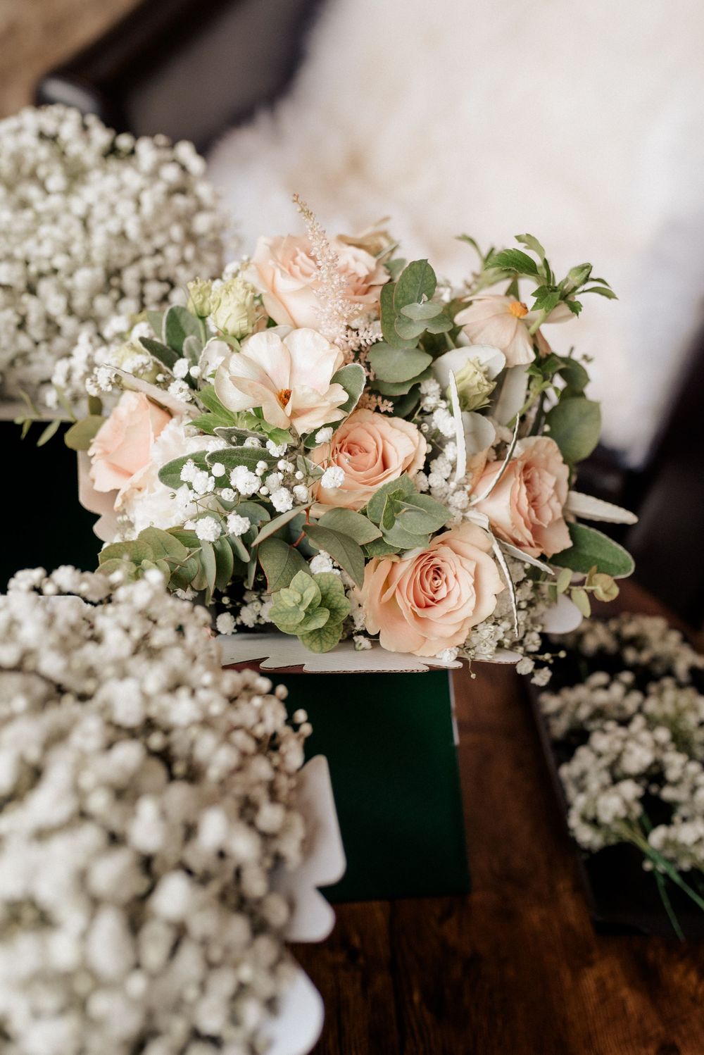 A delicate bridal bouquet of peach roses and baby's breath with eucalyptus leaves arranged on a dark wooden surface.