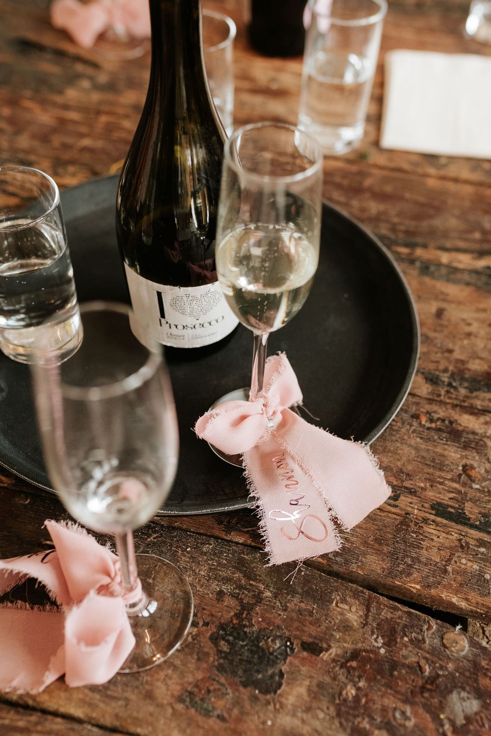 Wine glasses with delicate pink ribbons tied around the stems sit on a rustic wooden table.