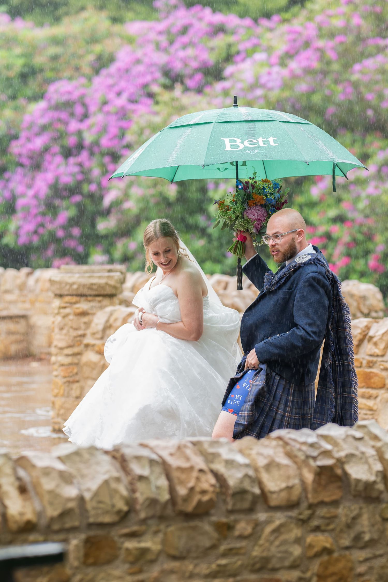 A couple shares a green umbrella on a stone wall with blooming pink azaleas in the background on their wedding day.