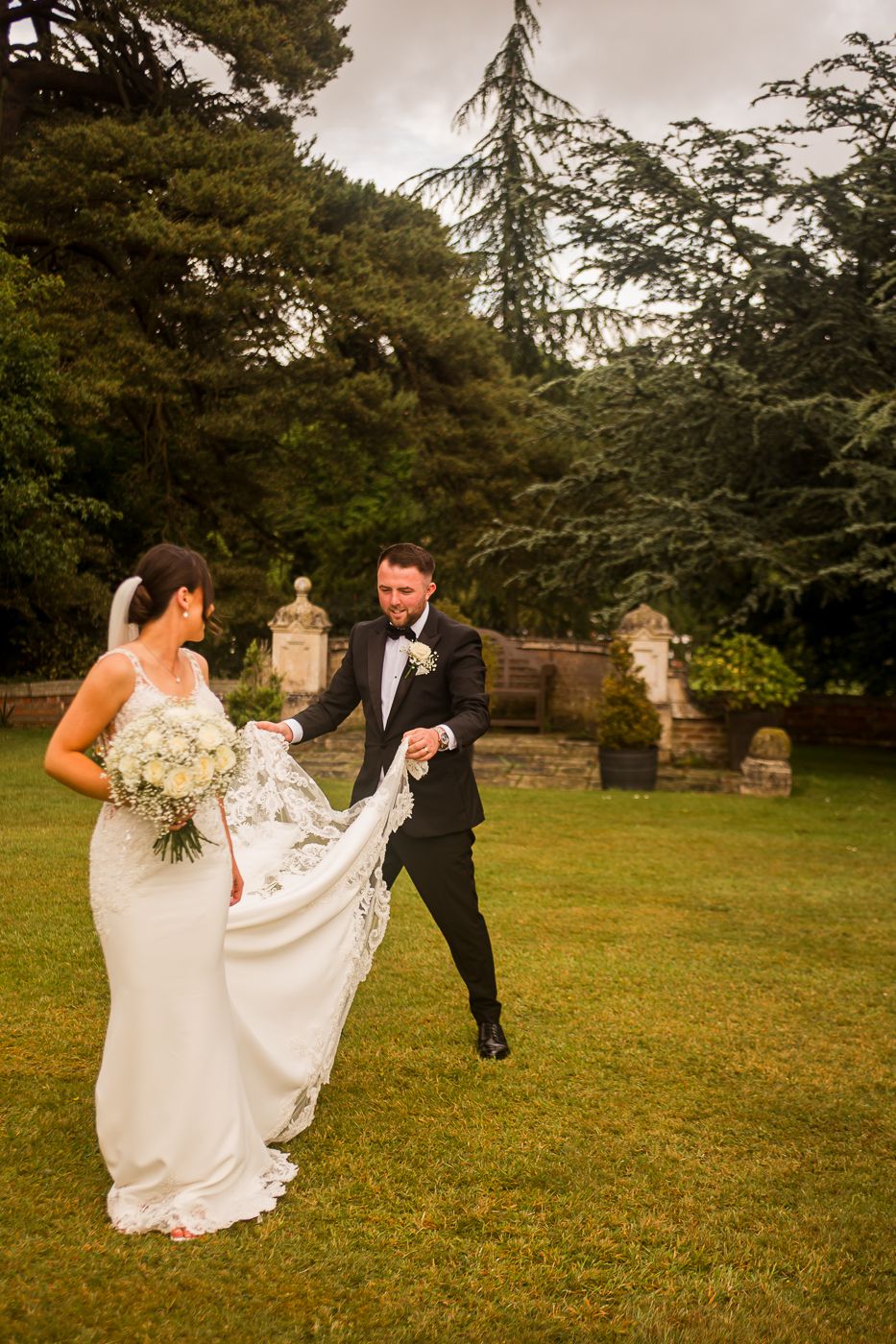 Newlyweds playfully walking and posing on a grassy lawn near evergreen trees.