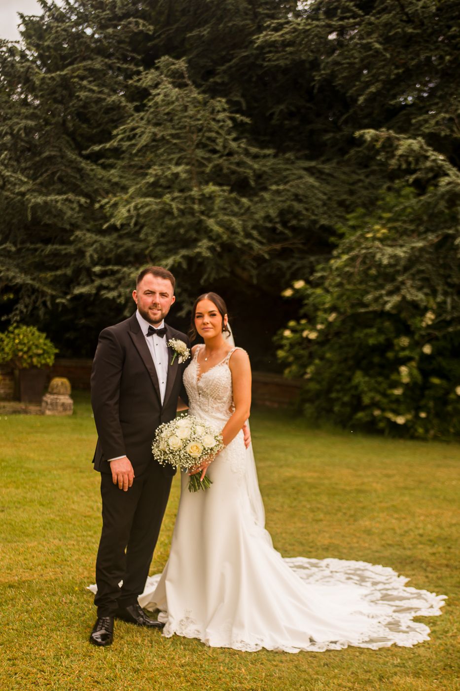 Wedding couple posing for portraits on a green lawn with trees in background.