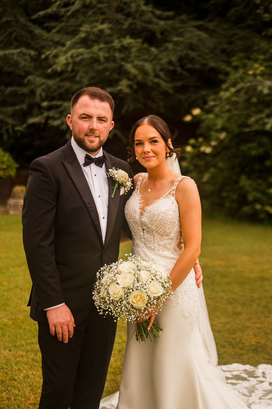 Newlyweds share a romantic moment on their wedding day in an outdoor garden setting with a white floral bouquet.