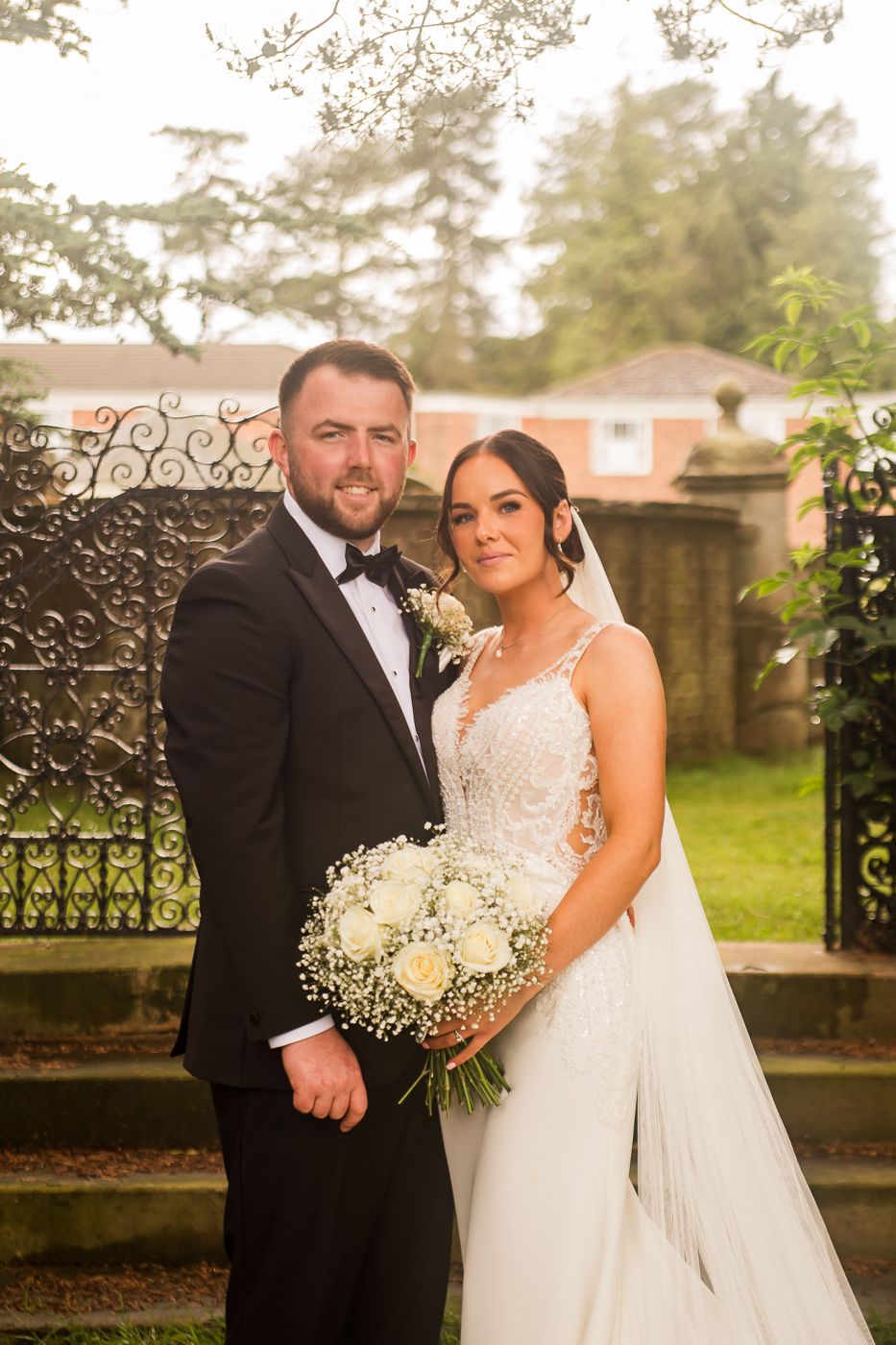 Wedding couple poses near decorative iron gate in garden setting with white floral bouquet.