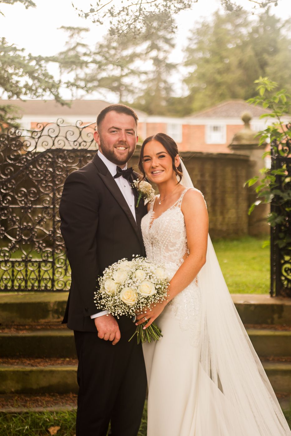 A romantic wedding couple poses by an ornate iron gate with a beautiful white rose and baby's breath bouquet.