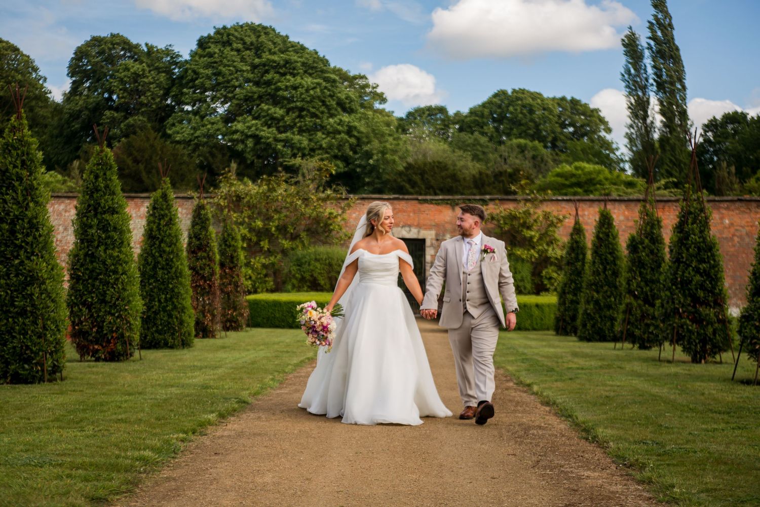 Wedding couple walks hand in hand through a manicured garden with cypress trees and brick walls.