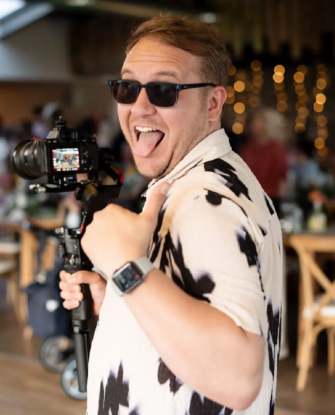 Photographer in a white shirt and sunglasses holds a camera while making a playful expression in a dimly lit venue.