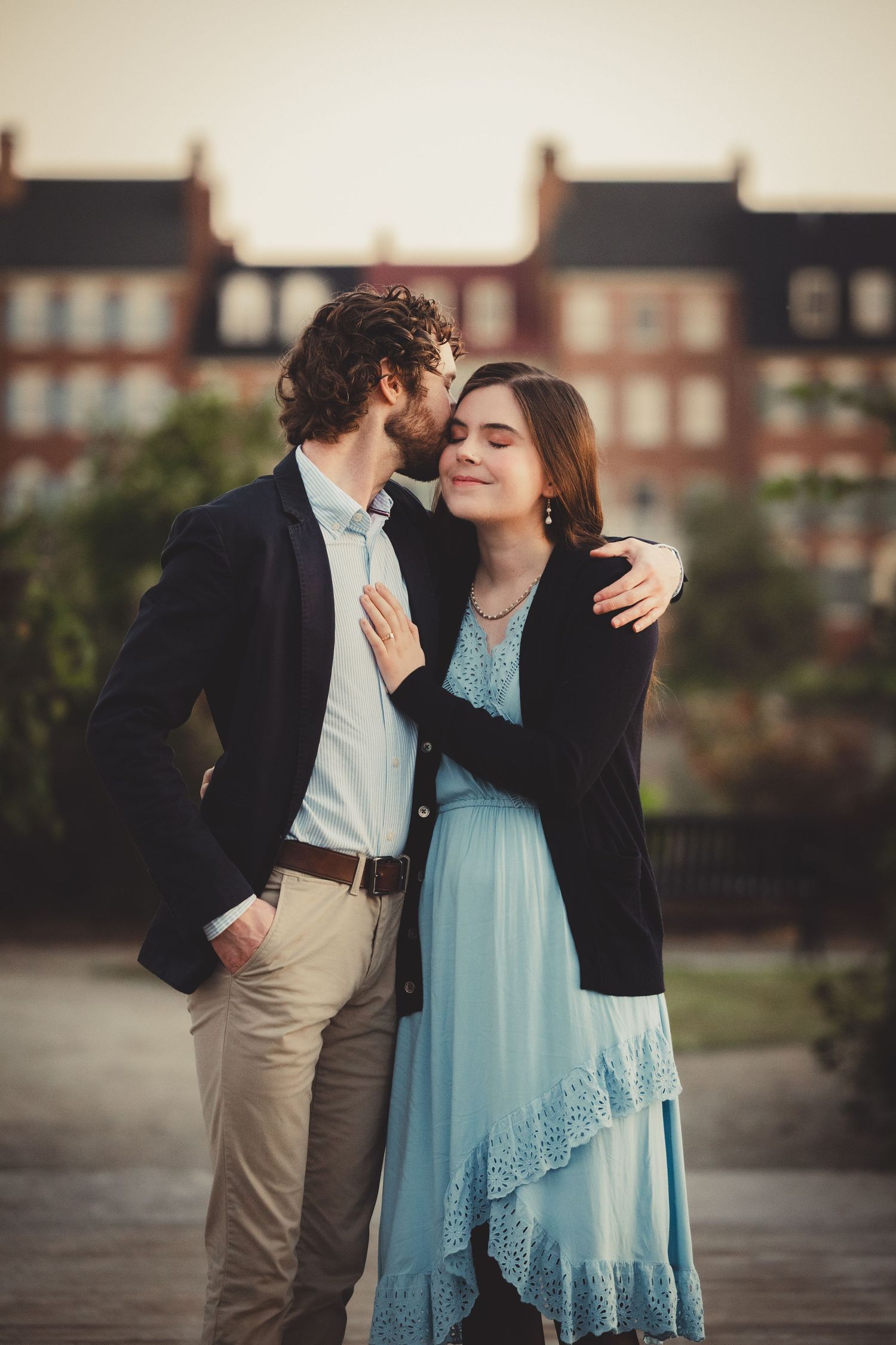 A romantic couple embraces outdoors, with the woman wearing a light blue dress and the man in a navy blazer.