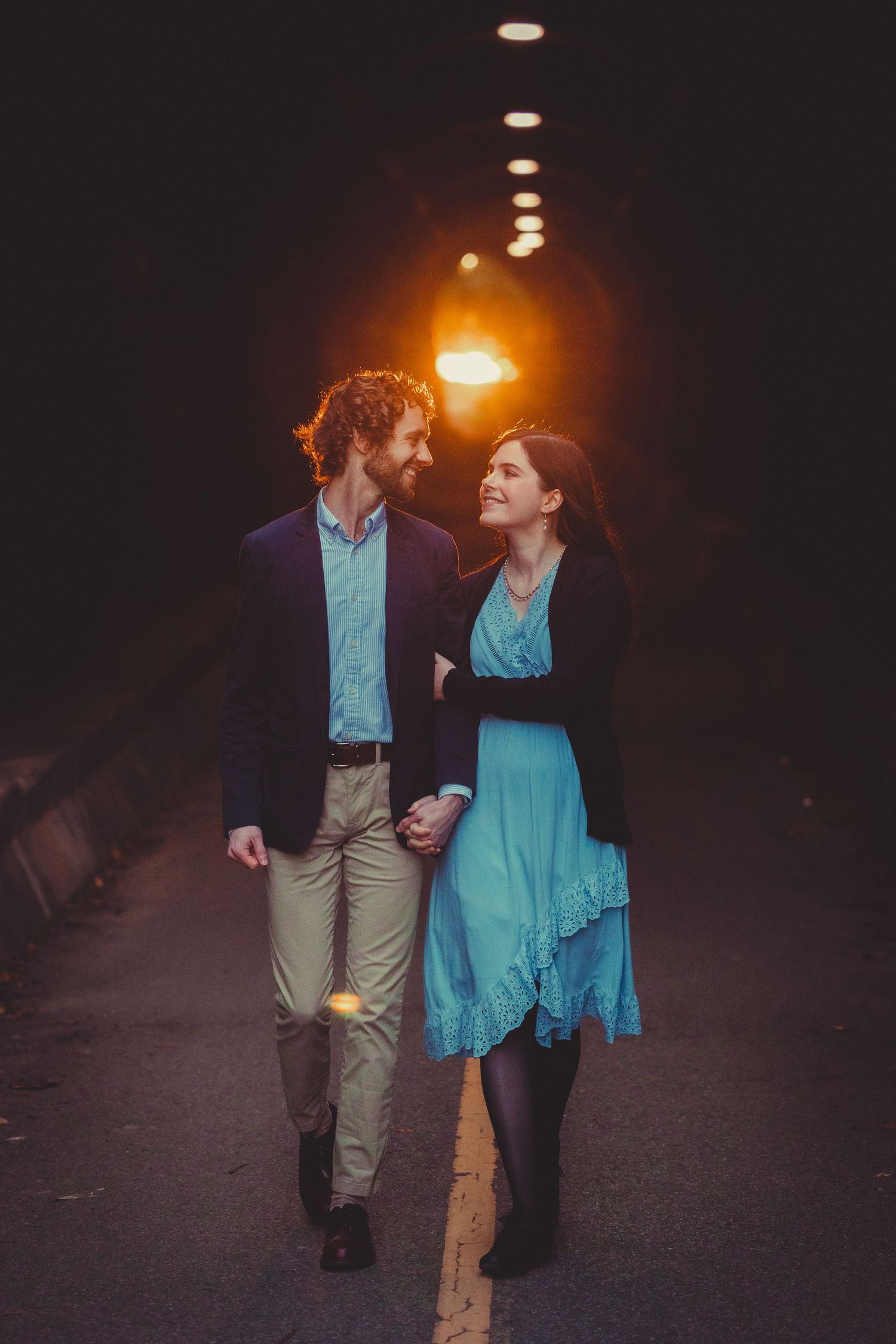 A couple in formal attire walks together under dim lighting, with the woman wearing a turquoise dress and man in navy blazer.