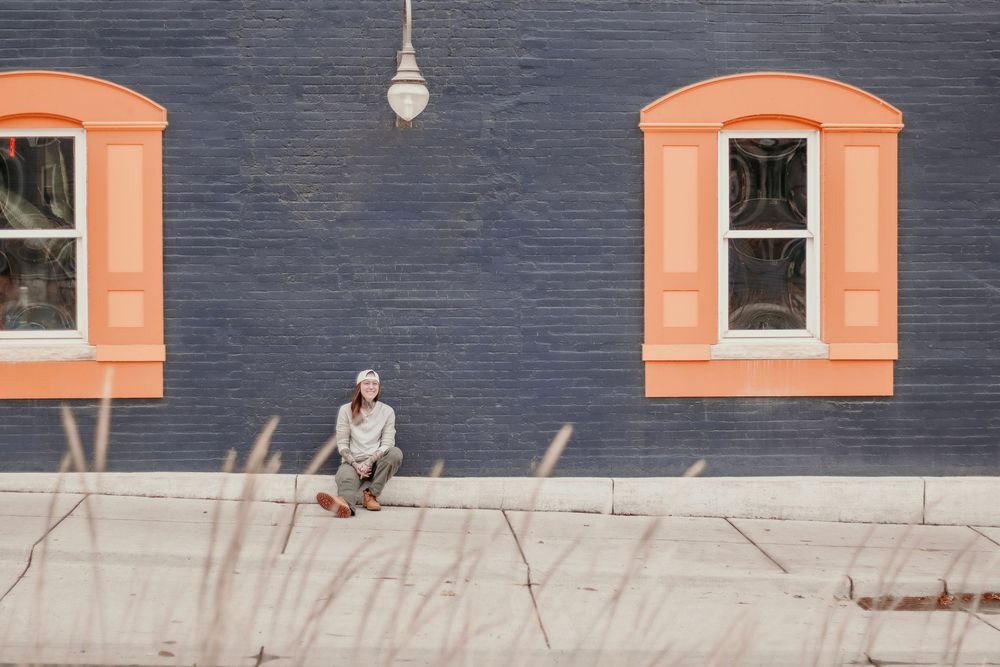 A woman sitting against a blue brick wall with orange shutters
