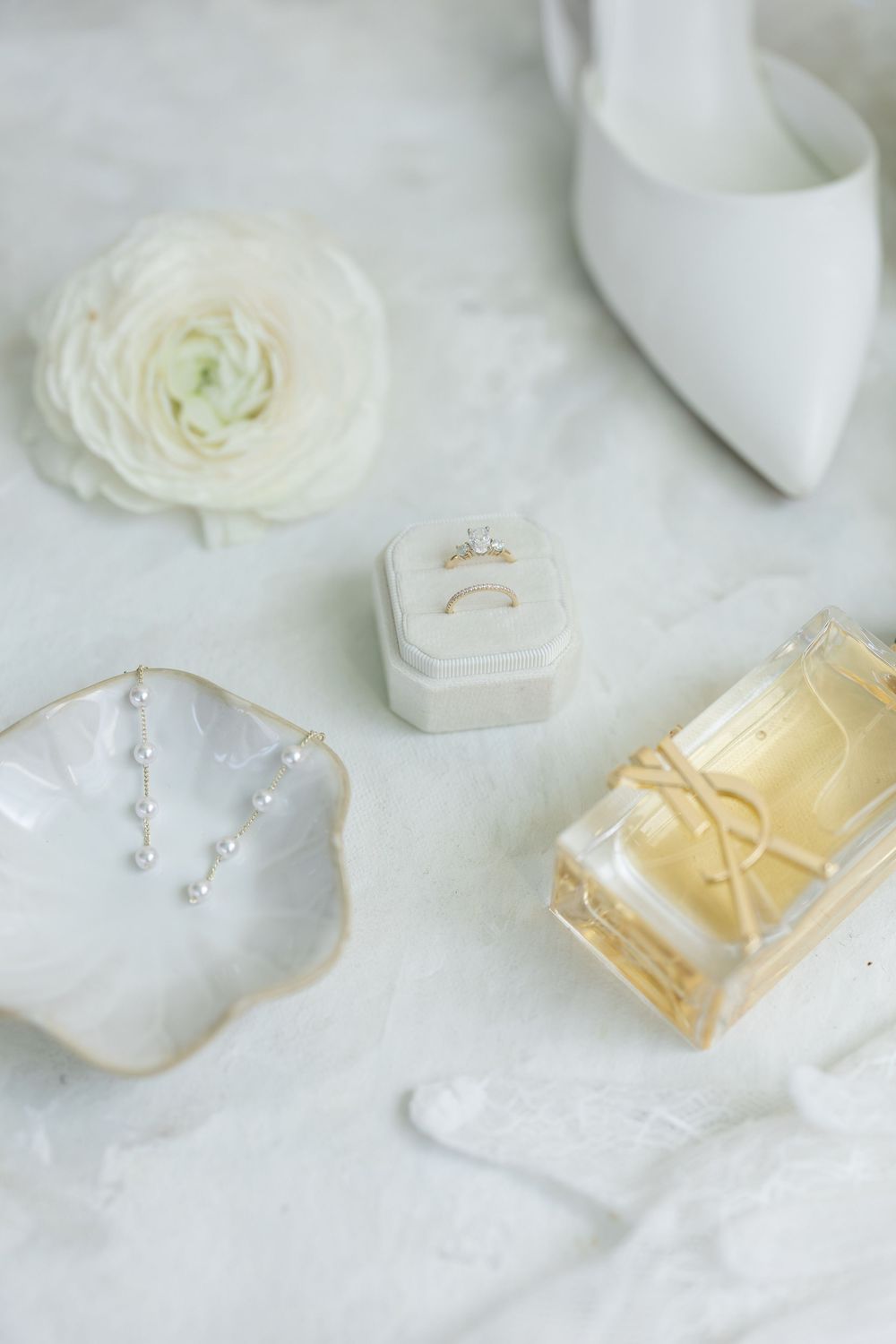 Wedding details featuring white roses, delicate rings, and jewelry on a light marble surface.