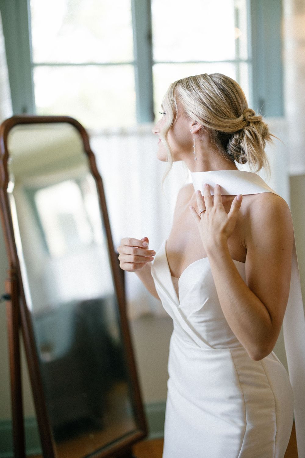 A bride in an elegant white gown looking at her reflection in a bright sunlit room.