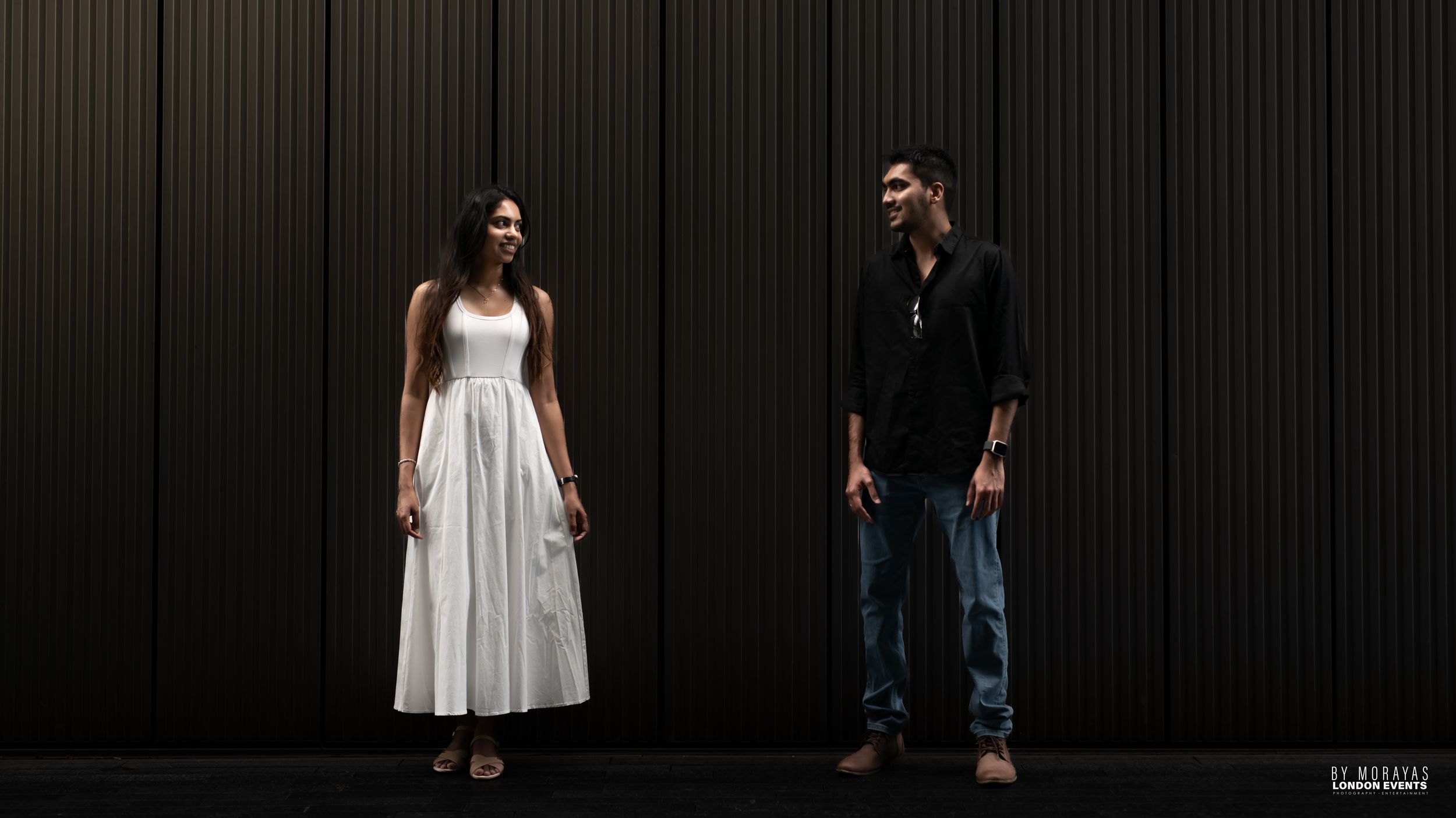 A couple poses against a dark vertical striped wall in contrasting black and white outfits.