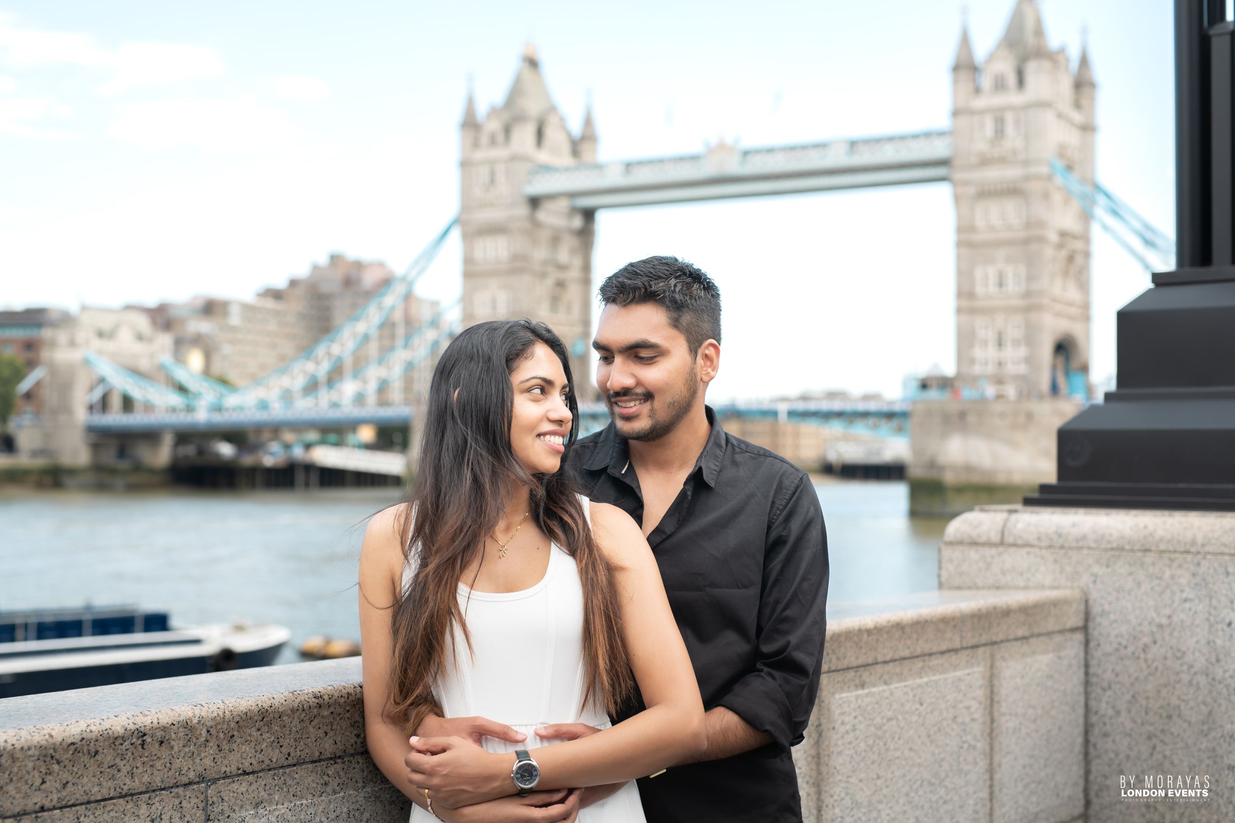 A couple embraces by the Thames River with Tower Bridge as their backdrop.