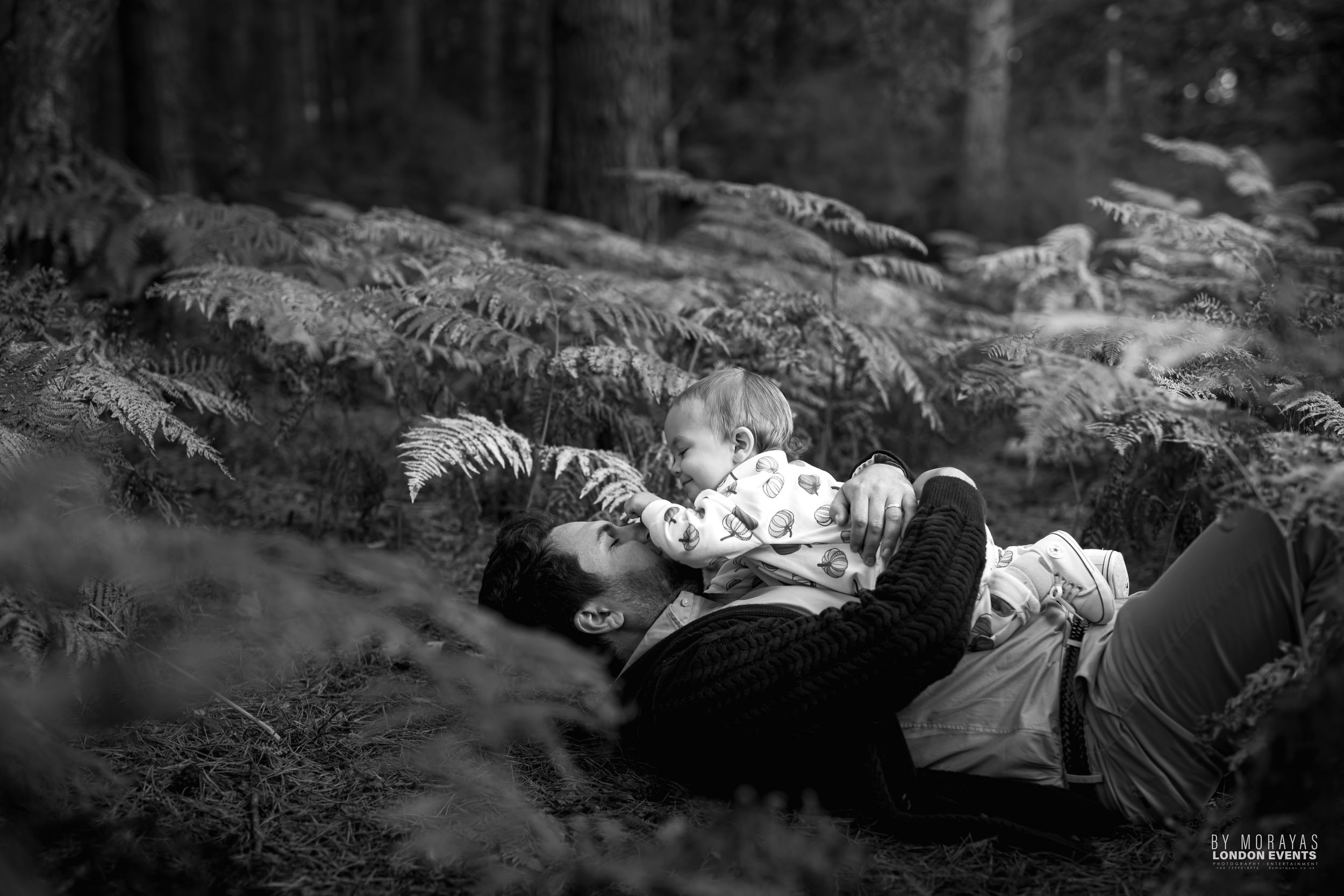 A black and white photo of people lying down together in ferns and grass in a forest setting.