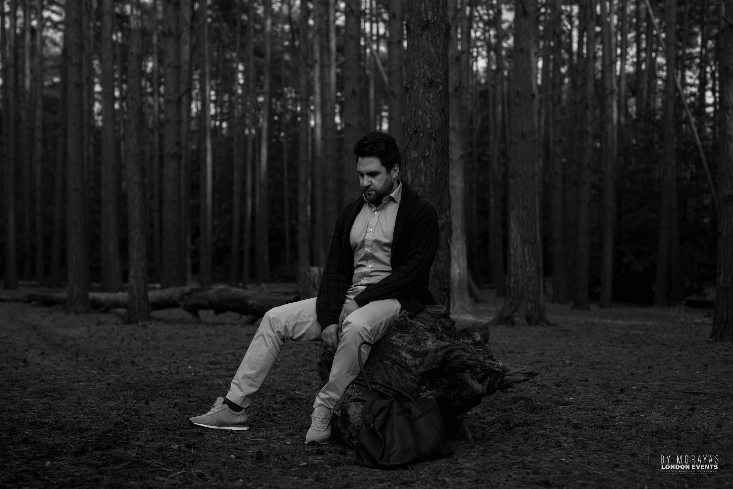 A black and white portrait of someone sitting on a log in a dark forest.