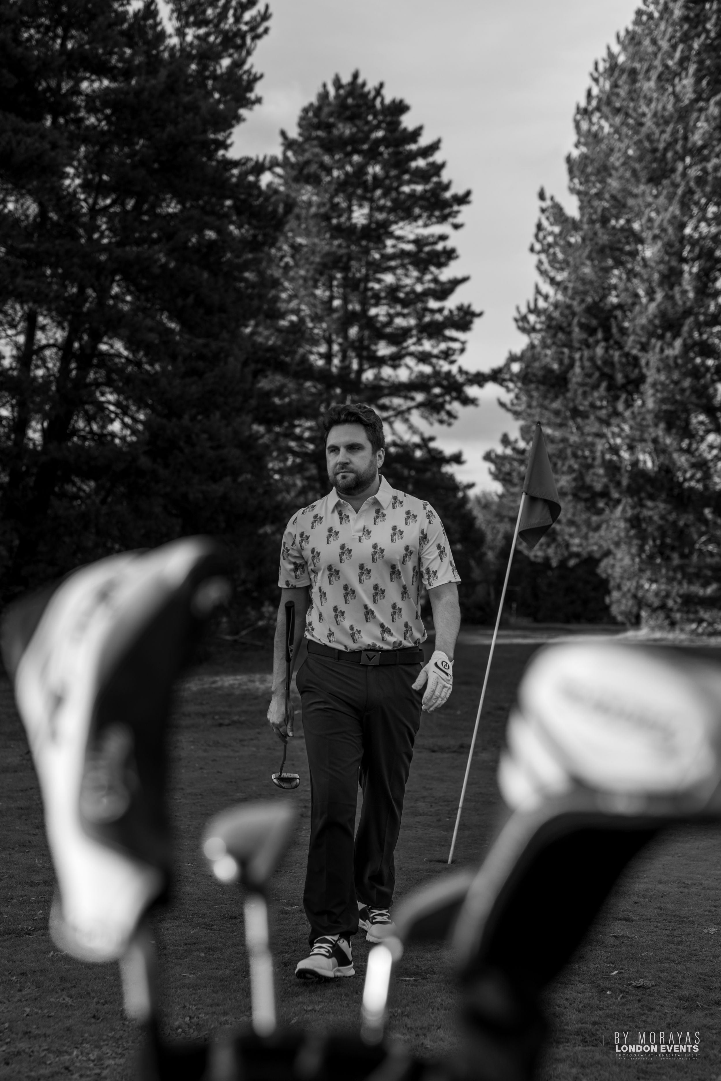 A golf scene captured through golf clubs in the foreground with pine trees in the background.