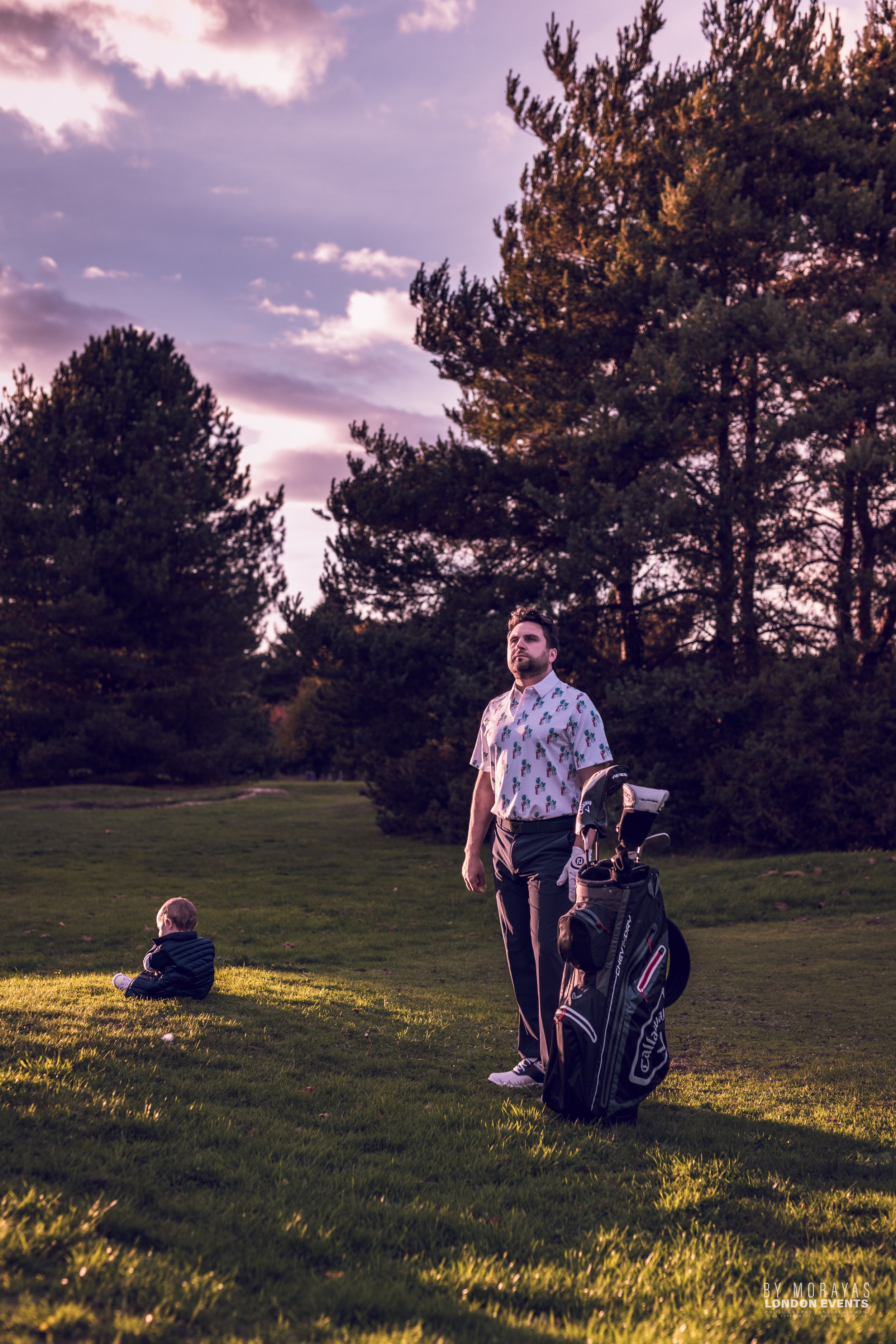 A golfer stands with golf bag on a course at sunset while a small child sits in the distance.
