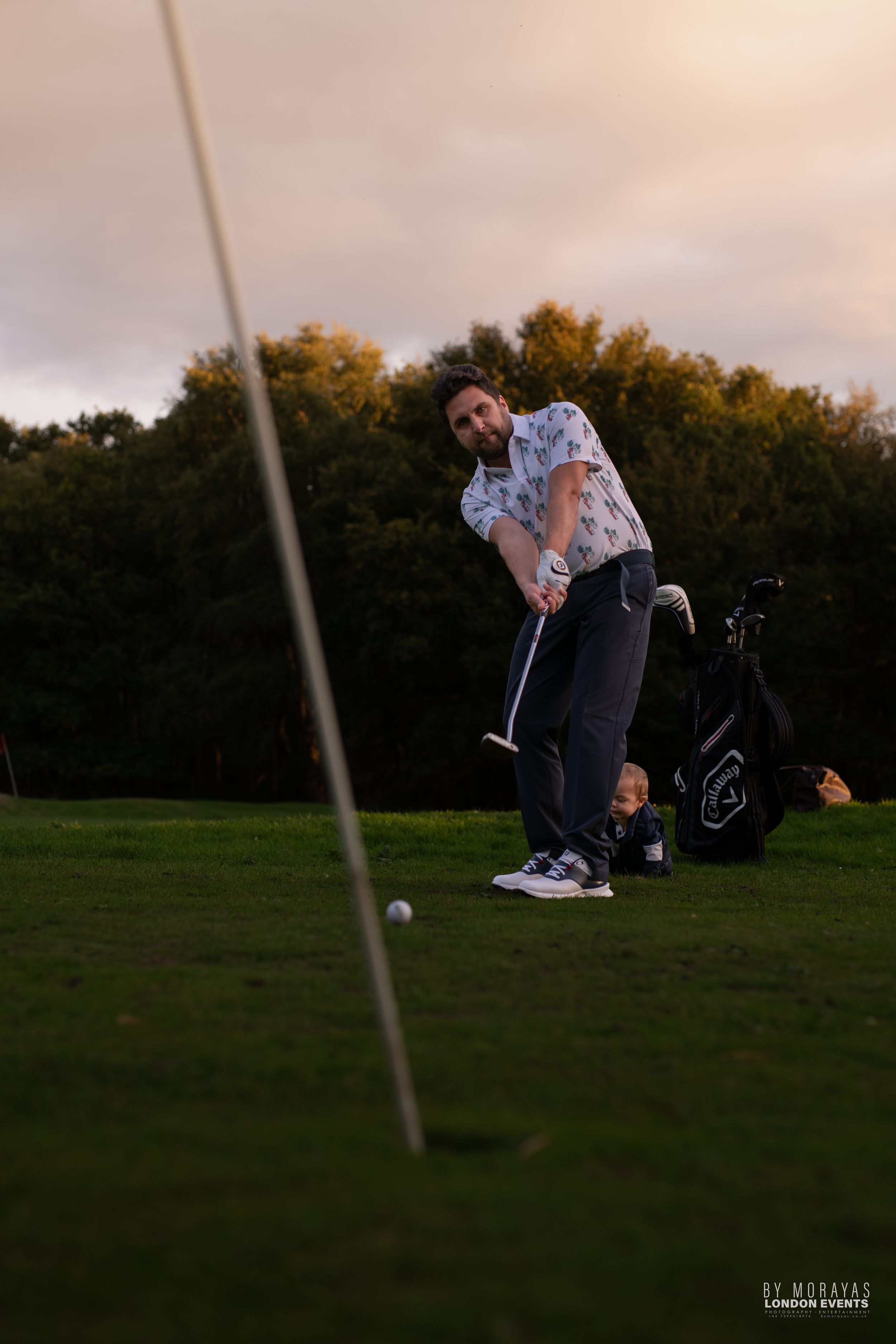 A golfer takes a swing on a golf course during golden hour with equipment visible.
