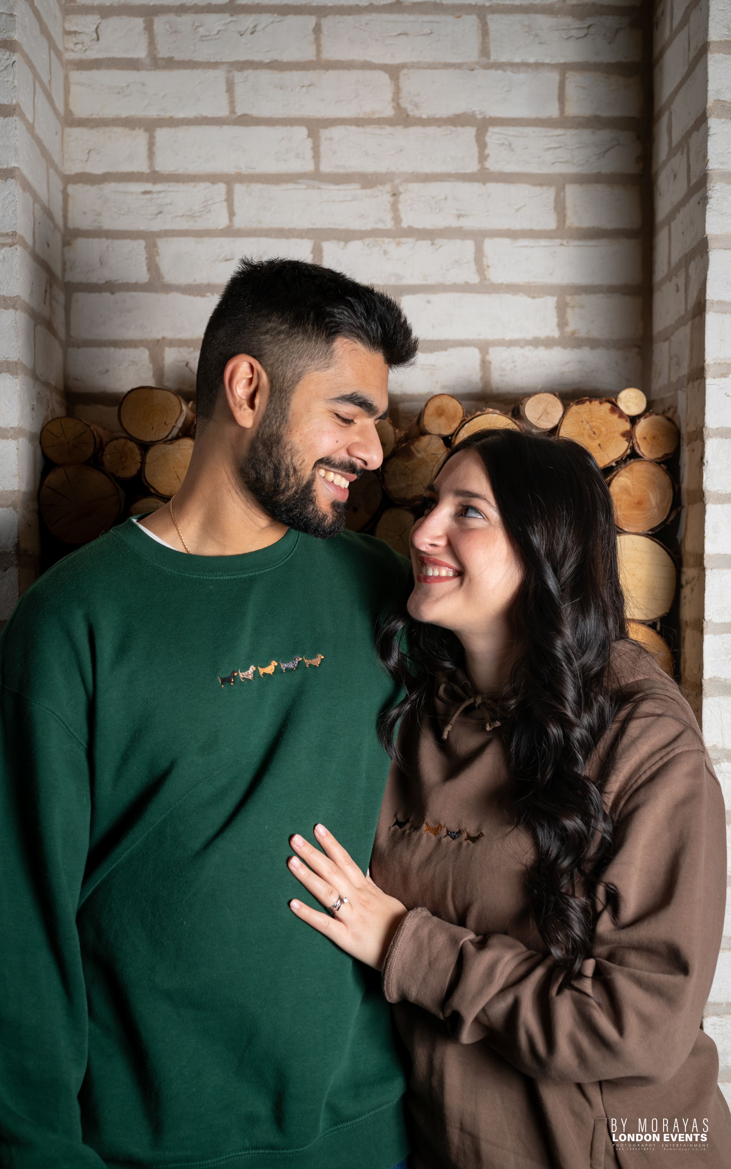A couple in green and brown sweaters share an intimate moment against a white brick wall with stacked firewood.
