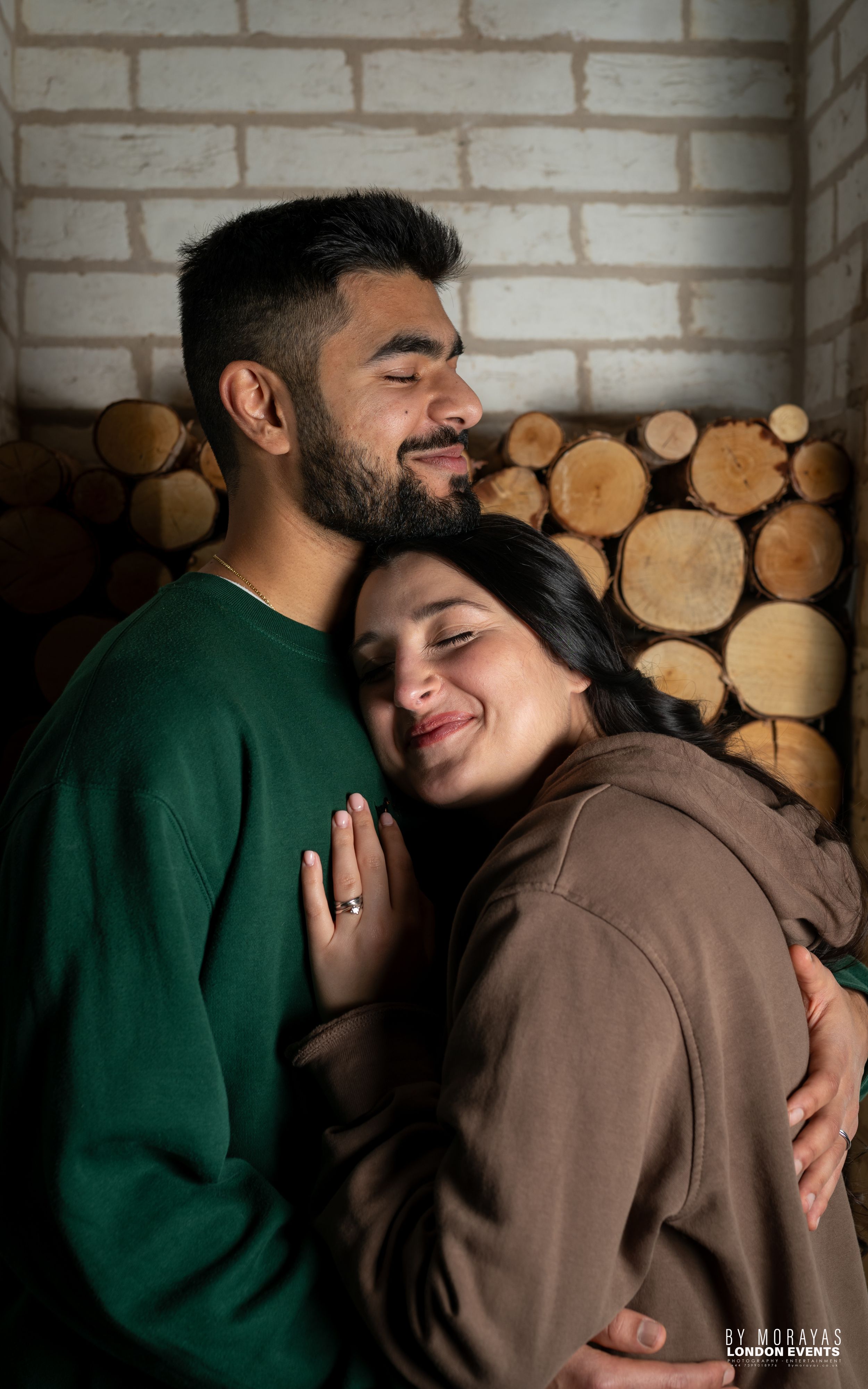 An affectionate couple in casual sweaters embraces against a rustic brick wall backdrop.
