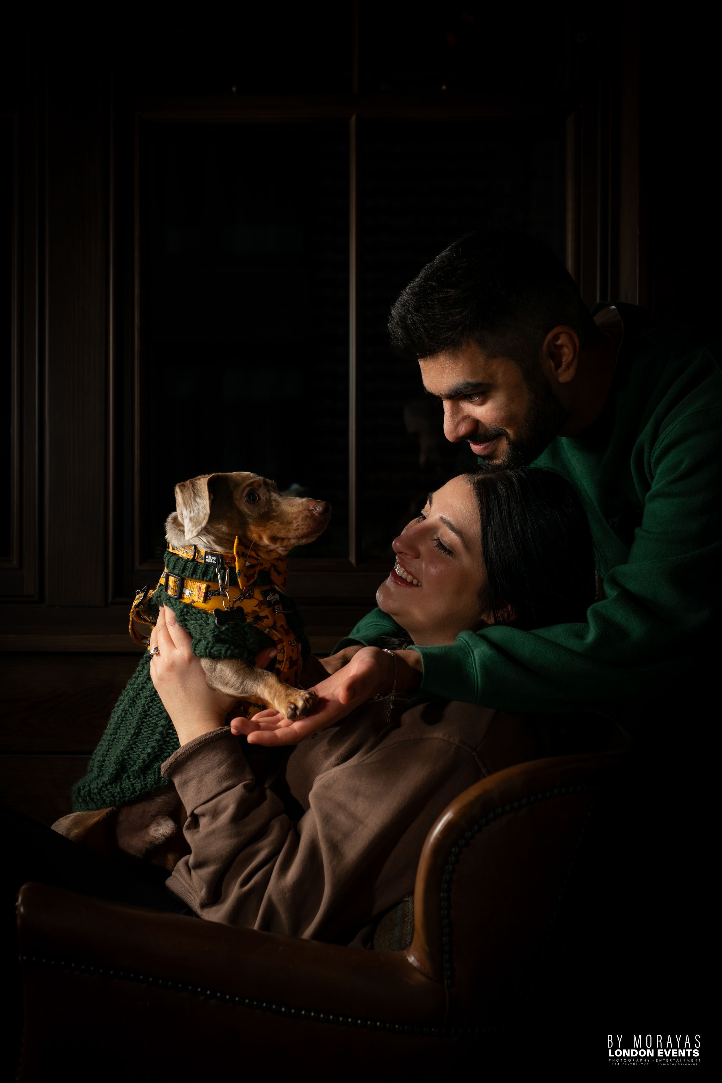 A couple and their dog share a cozy moment on a leather couch in low-key lighting.