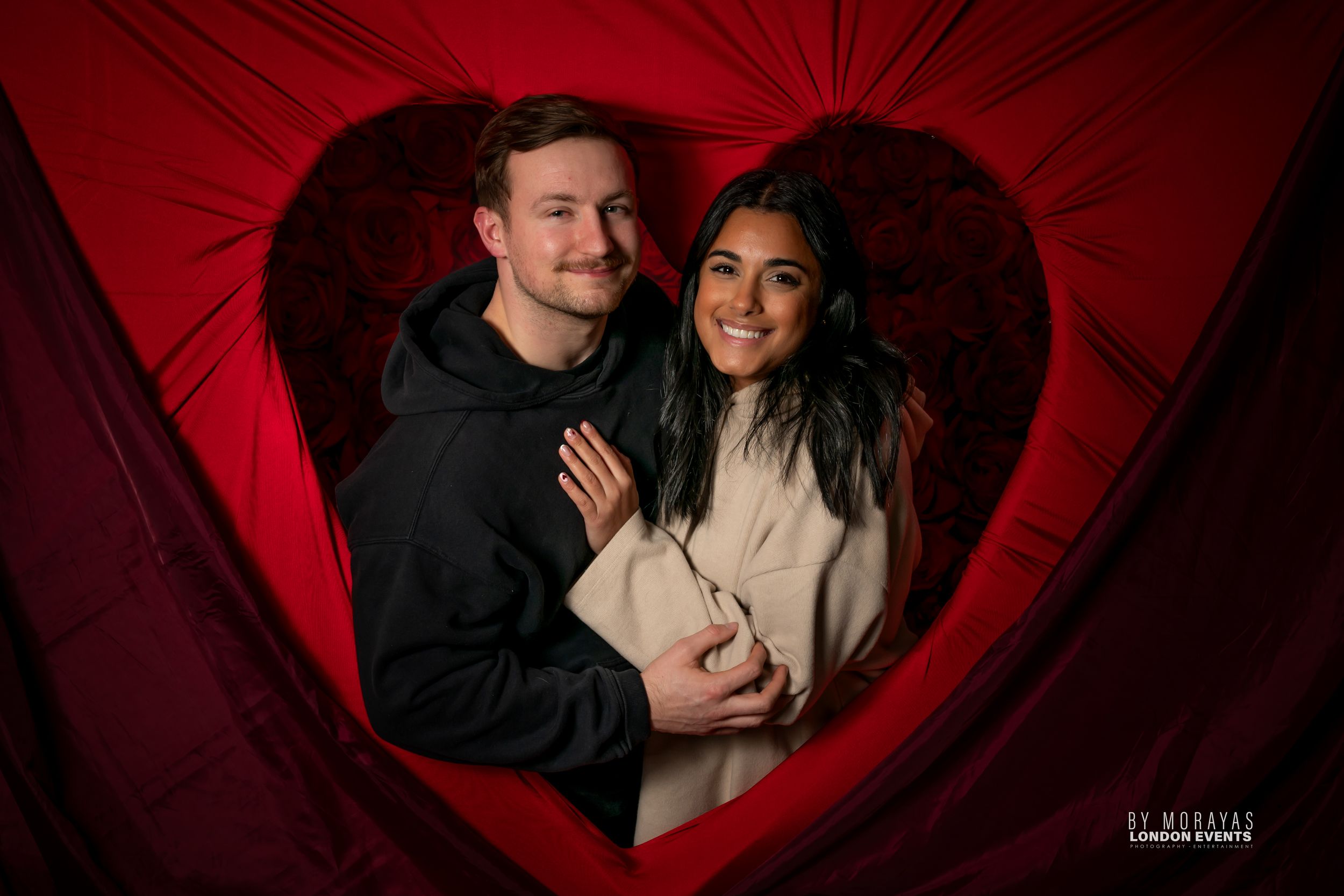 Romantic couple posing against red heart-shaped backdrop with dramatic lighting.