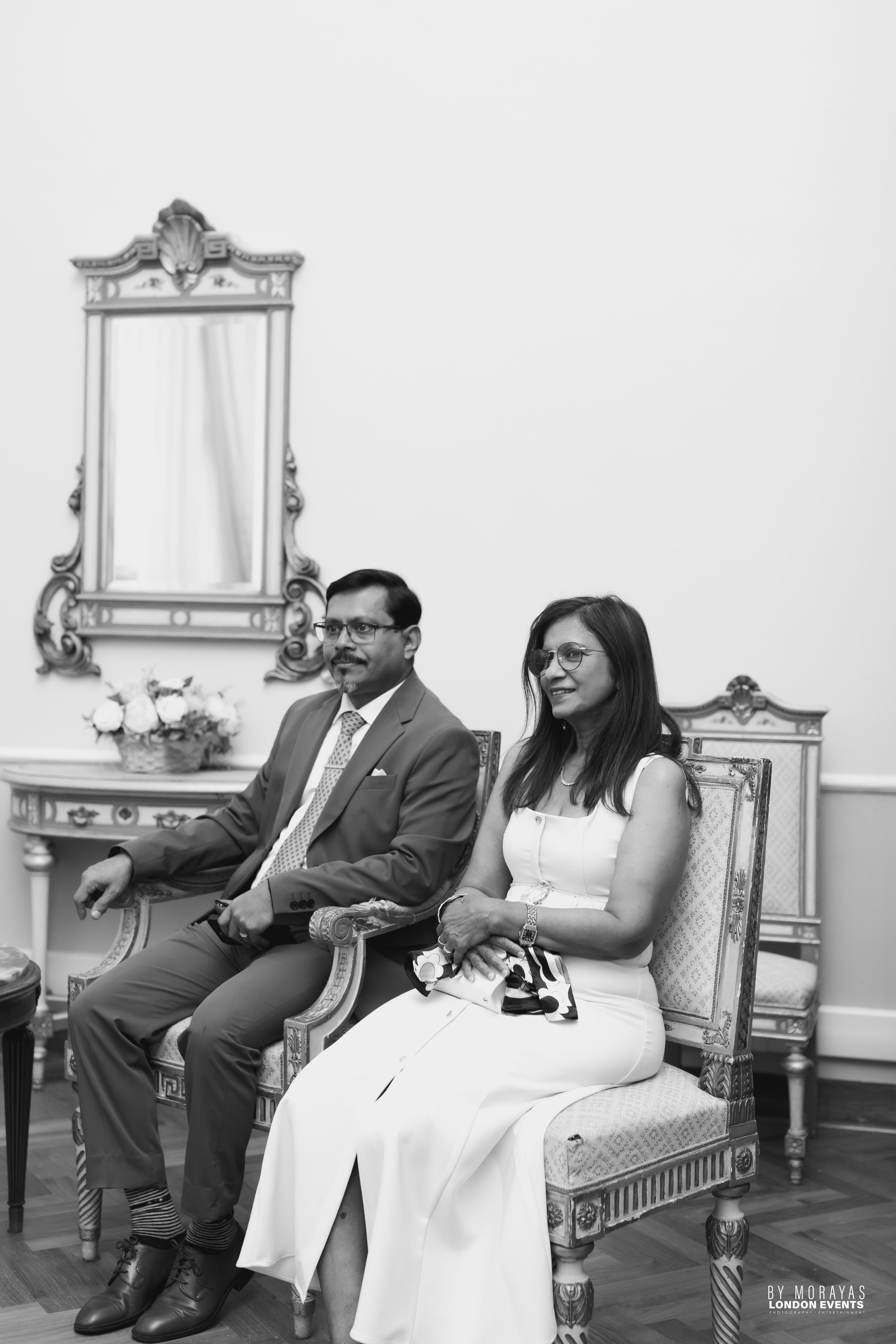 A couple relaxes on ornate white chairs during an indoor photoshoot.