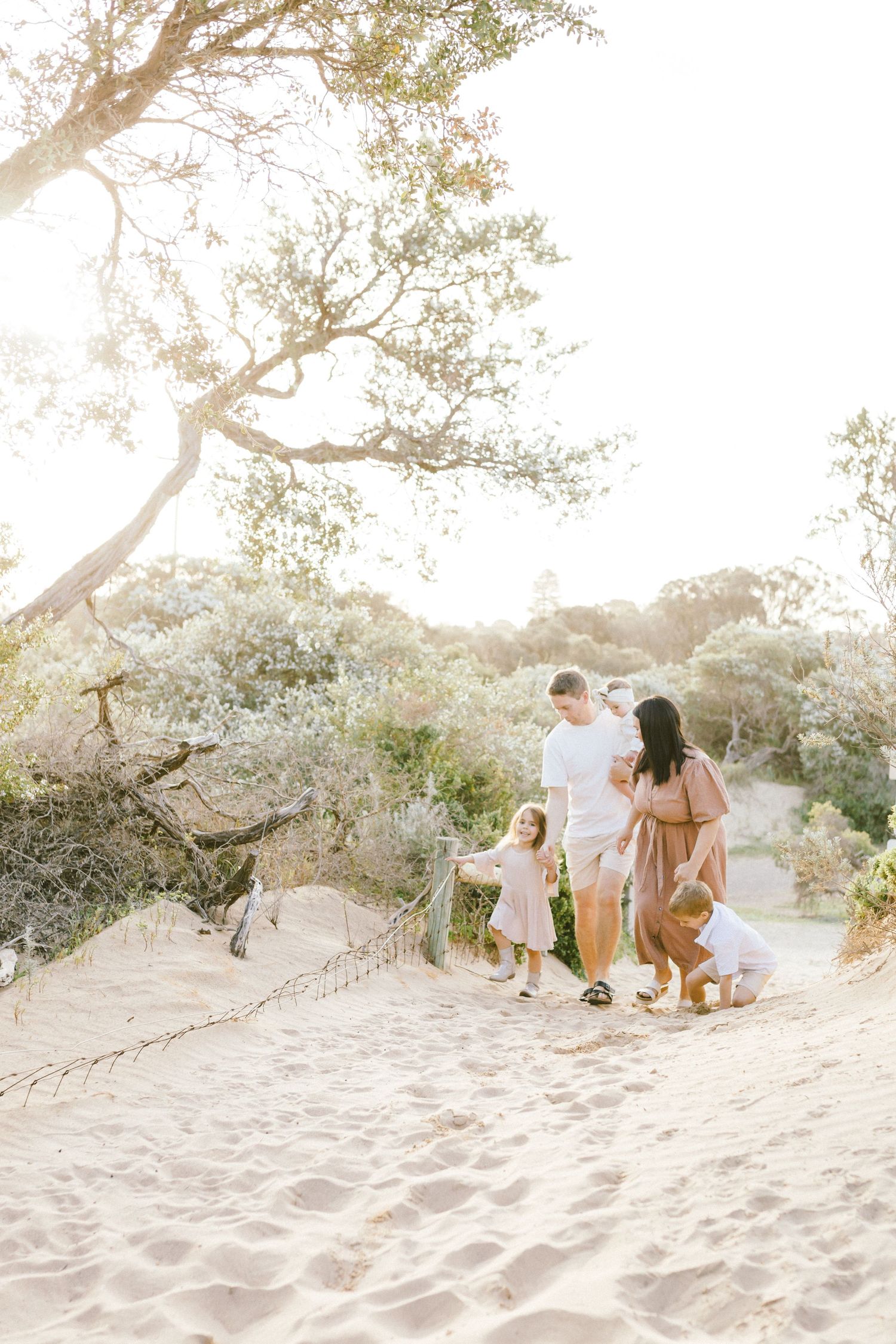 A family enjoys a tender moment together on a sandy beach path surrounded by coastal trees in soft golden light.