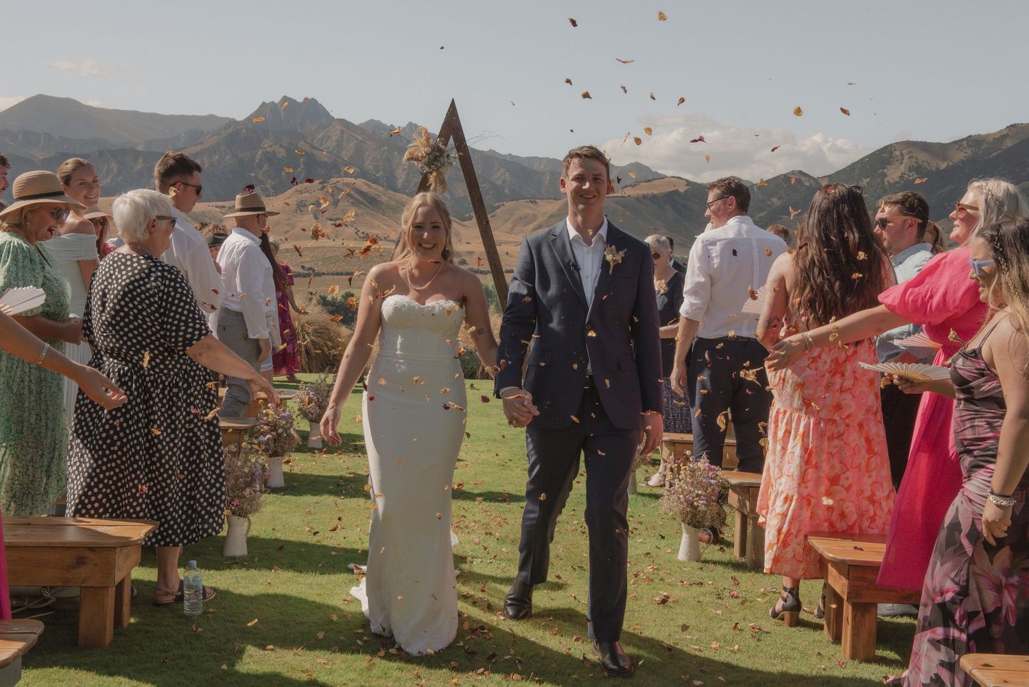 Guests throw flower petals at an outdoor desert wedding ceremony with mountain peaks in the background.