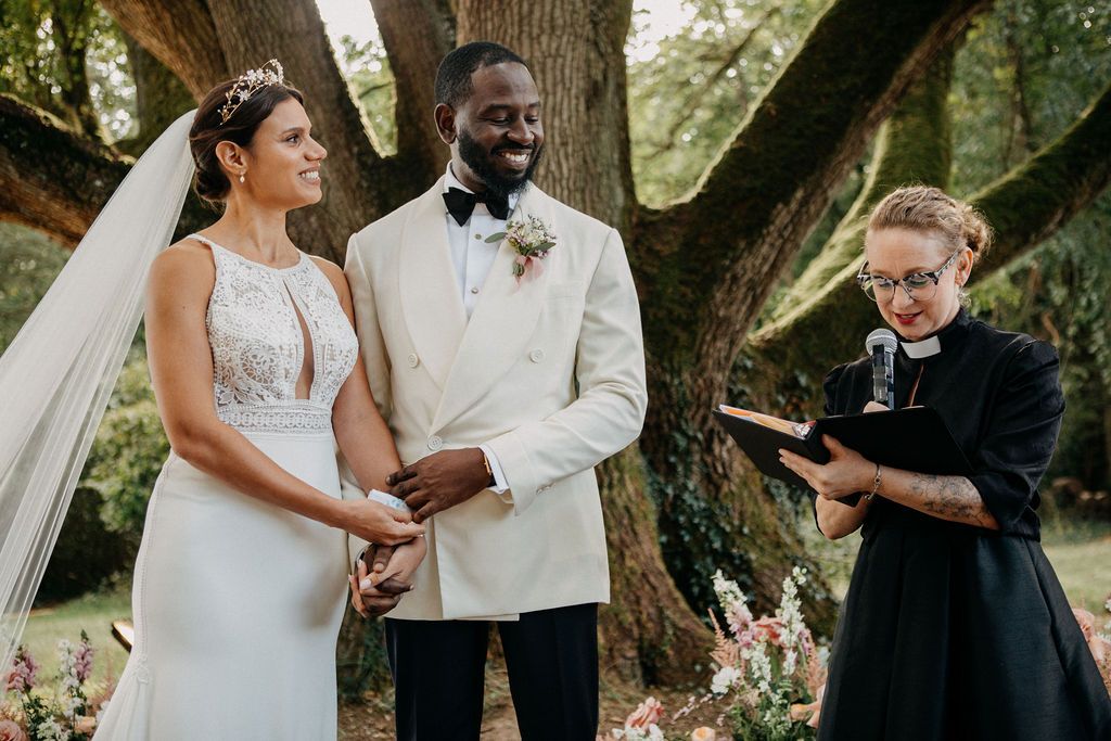 Bride and groom holding hands during outdoor wedding ceremony with officiant reading from book.