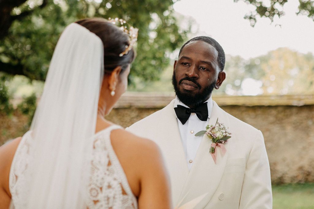 Groom in white tuxedo with boutonniere gazing at bride during outdoor wedding ceremony.