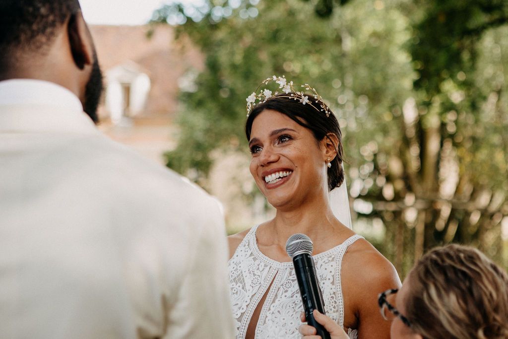 Smiling bride holding microphone during outdoor wedding ceremony wearing white dress and floral crown.