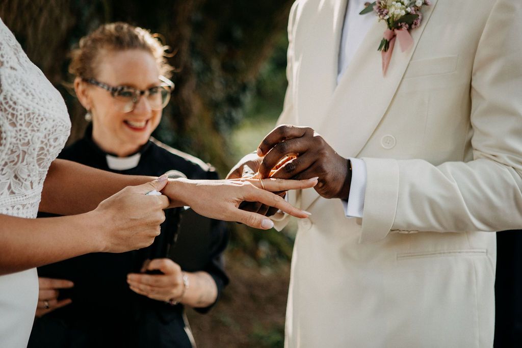 Couple exchanges rings during outdoor wedding ceremony as smiling officiant watches closely.