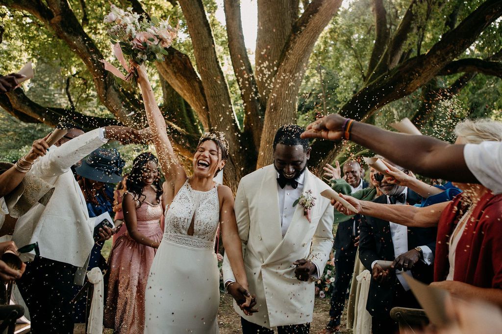 Newlyweds celebrating with confetti toss surrounded by cheering guests under large tree.