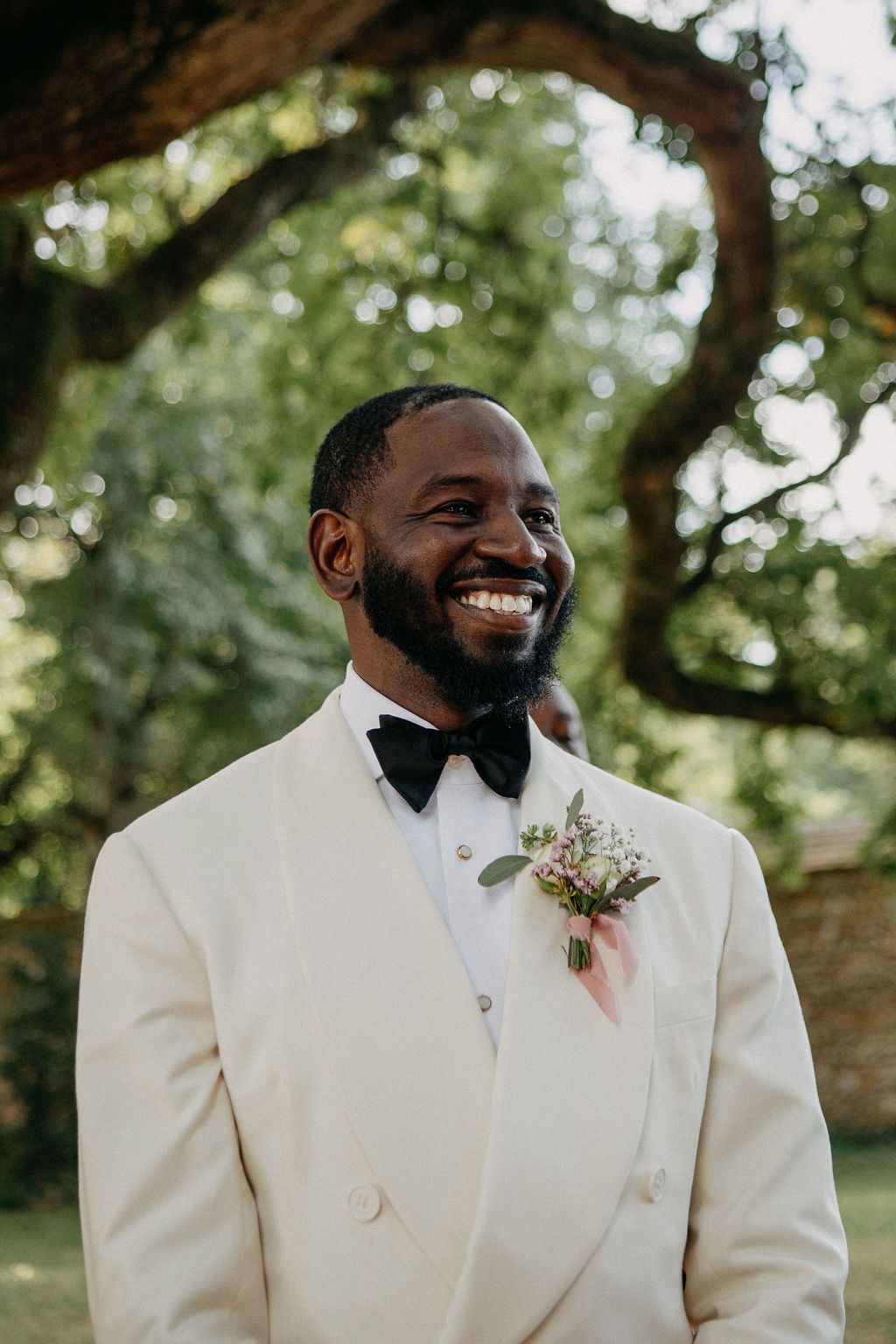 Smiling groom in white double-breasted tuxedo with boutonniere stands outdoors under green trees.
