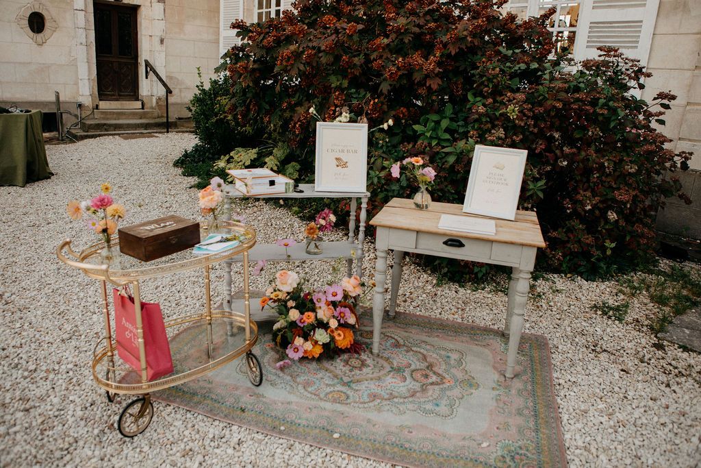Outdoor wedding welcome display with vintage bar cart, floral arrangements, and signage on gravel.