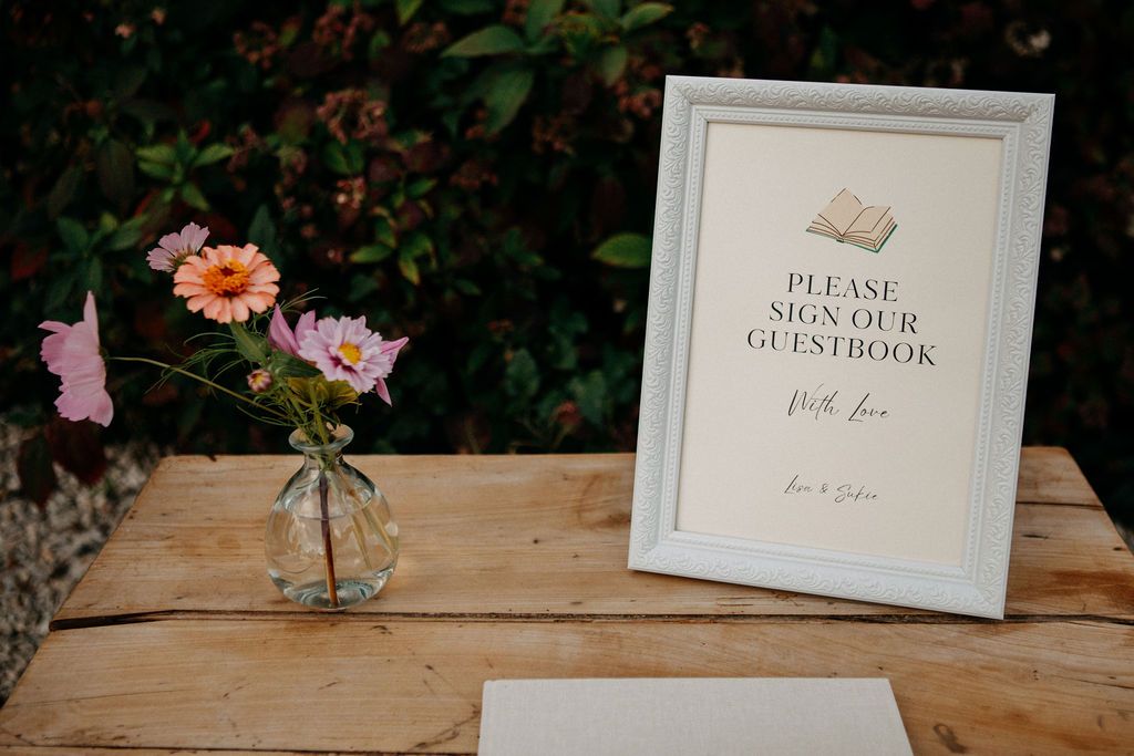 Wedding guestbook sign with small floral arrangement on rustic wooden table.