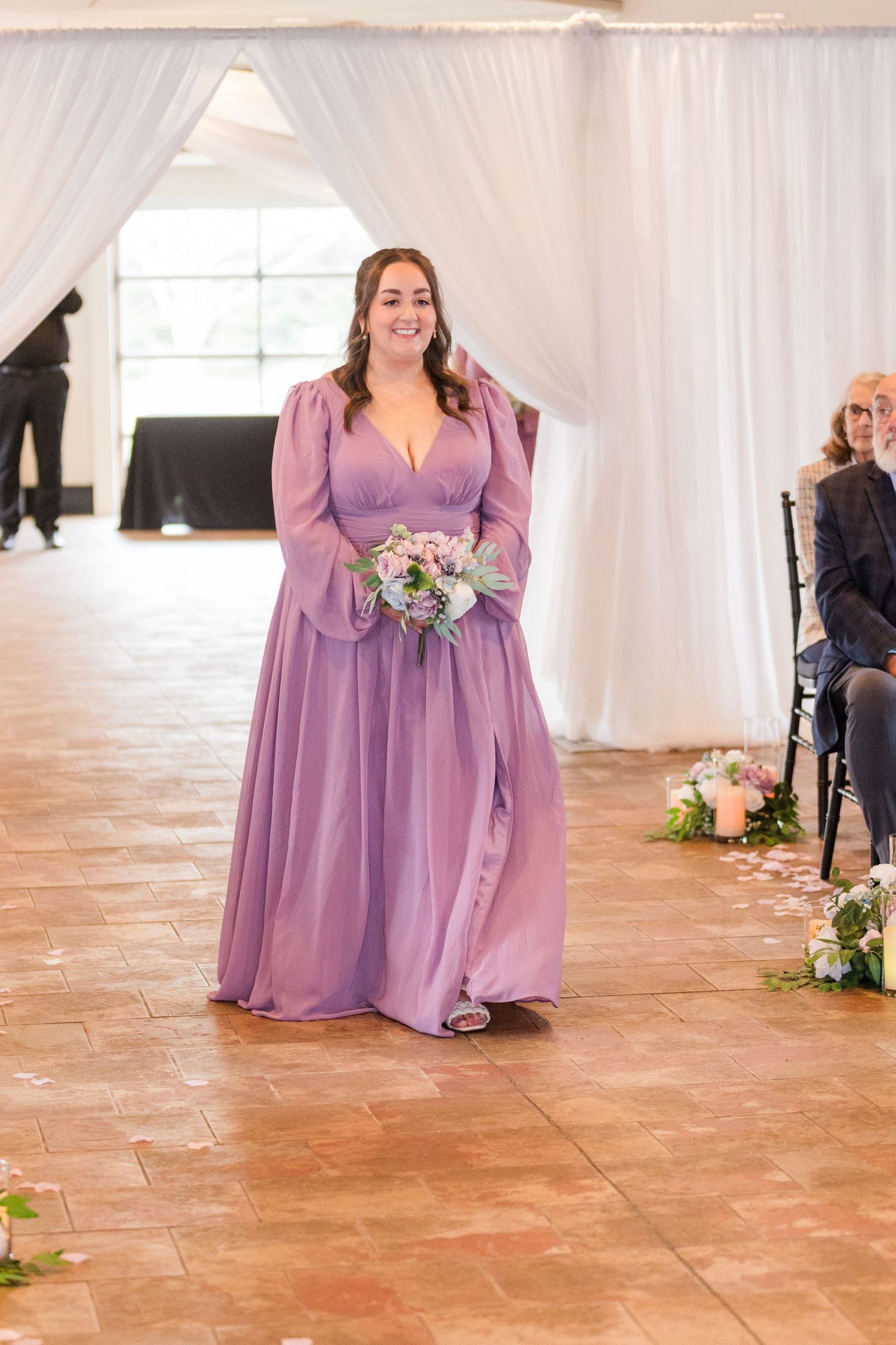 A bridesmaid in a flowing lilac purple dress walks down the aisle at an elegant indoor wedding ceremony.