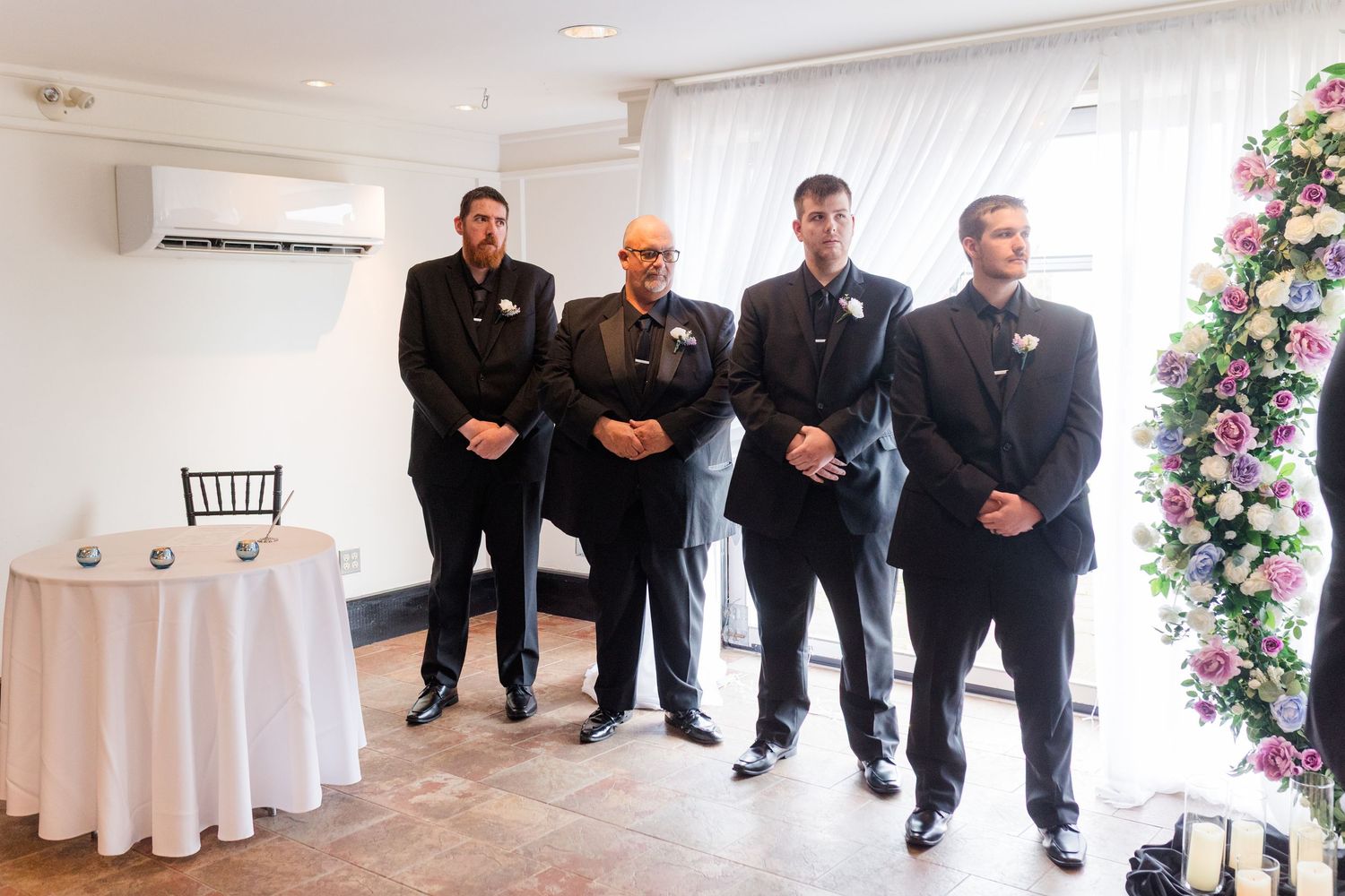 Four men in black suits stand together at a wedding venue with a white tablecloth and floral arrangement.