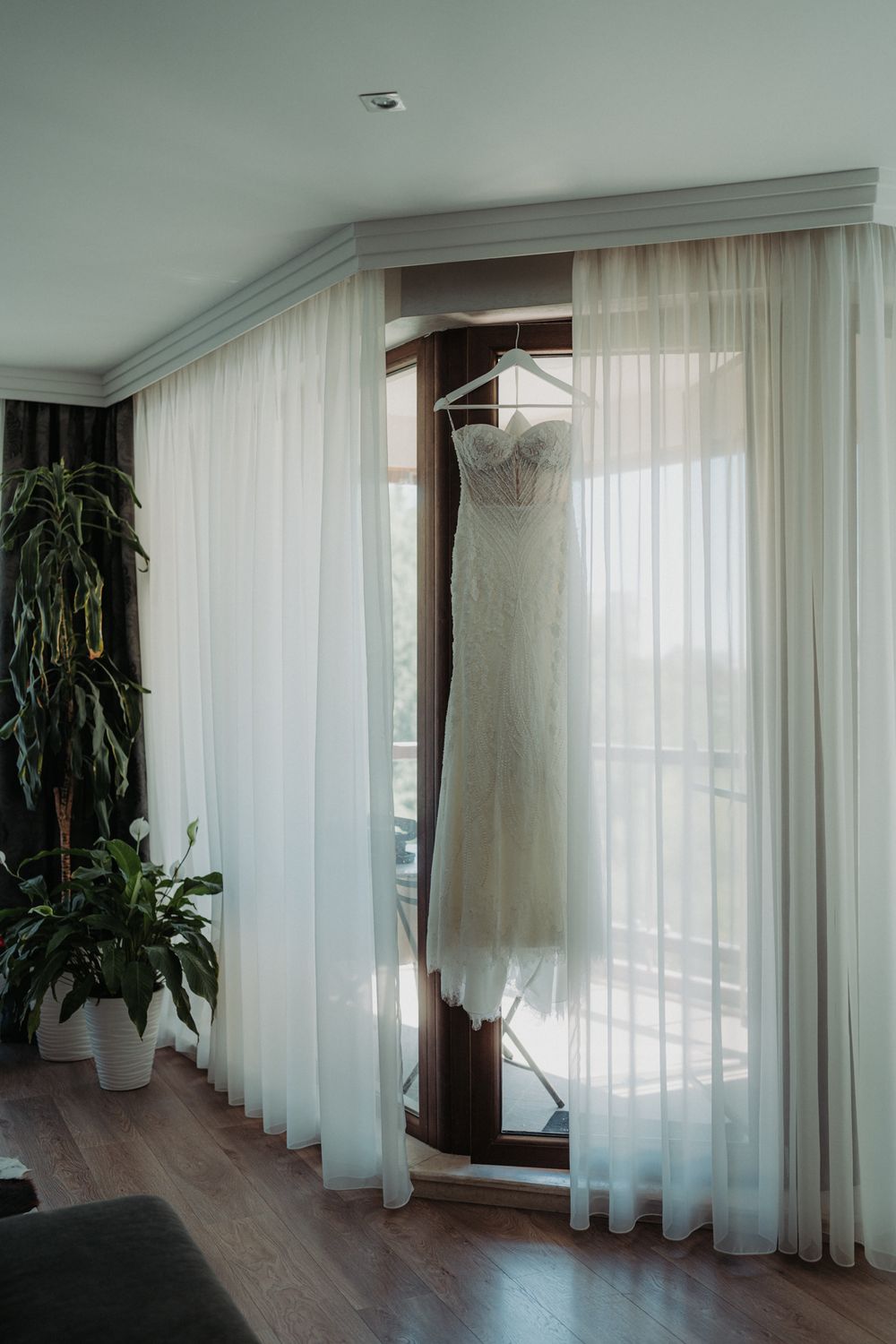 Elegant white wedding dress hanging in window with sheer curtains and indoor plants.