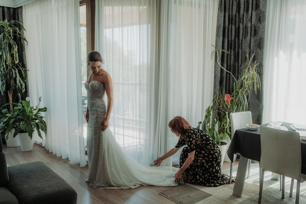A bride in a fitted white gown gets her dress adjusted in a bright room with sheer curtains and indoor plants.