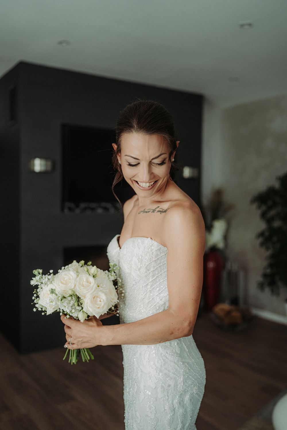 A bride in a strapless lace wedding dress holds a white floral bouquet while looking down.