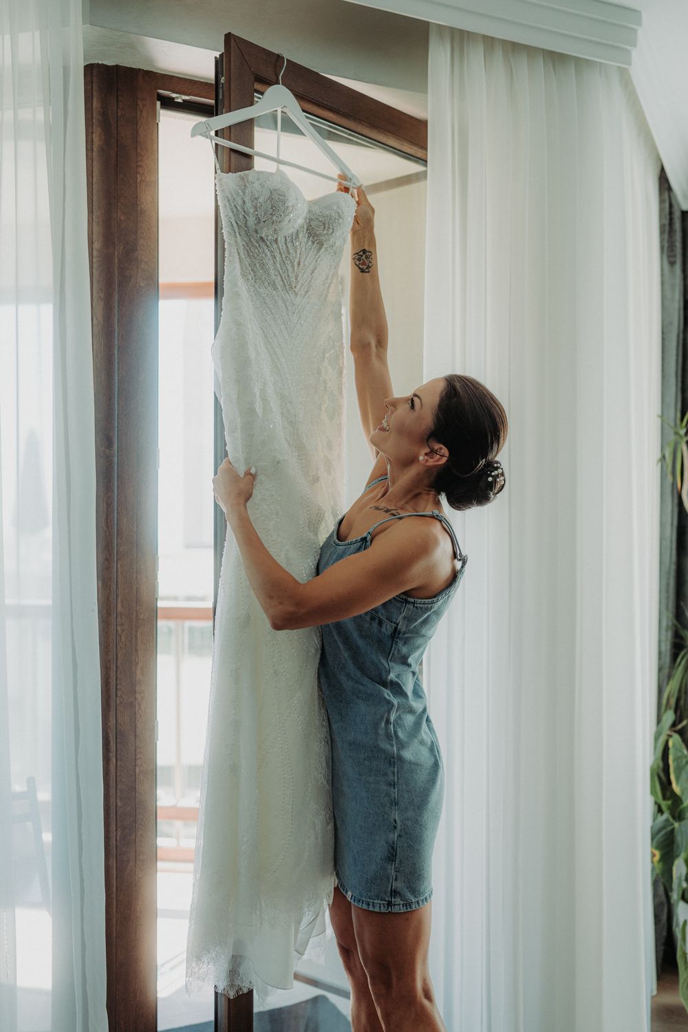 Person in denim adjusting a white wedding dress hanging by a window with sheer curtains.