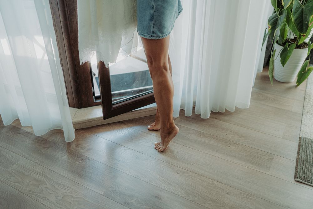 Bare feet standing on light wooden flooring next to white curtains.