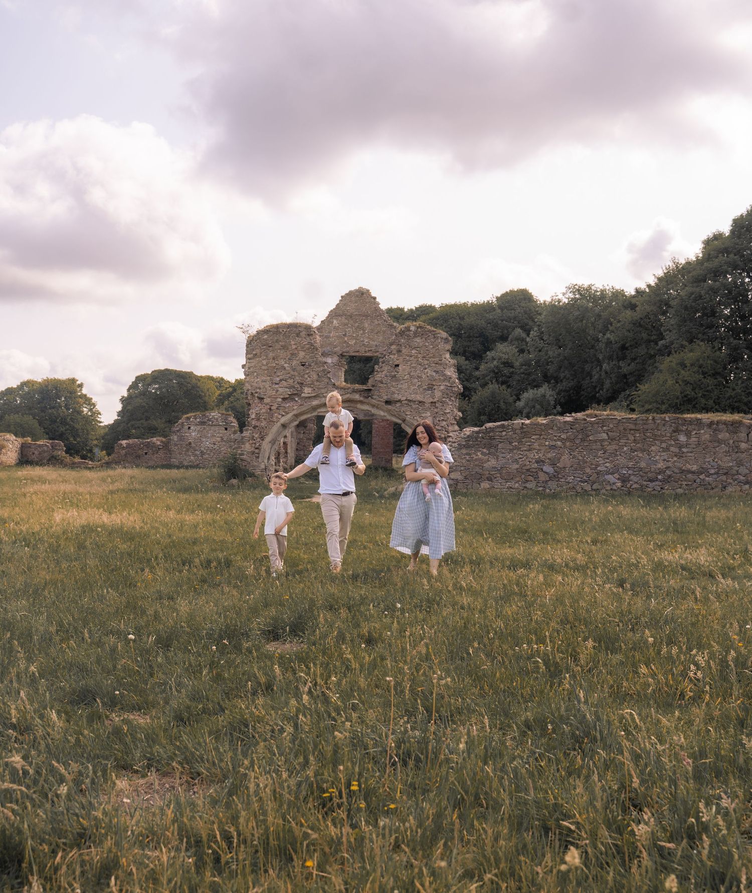 A family walks through a grassy field near ancient stone ruins on a cloudy summer evening.