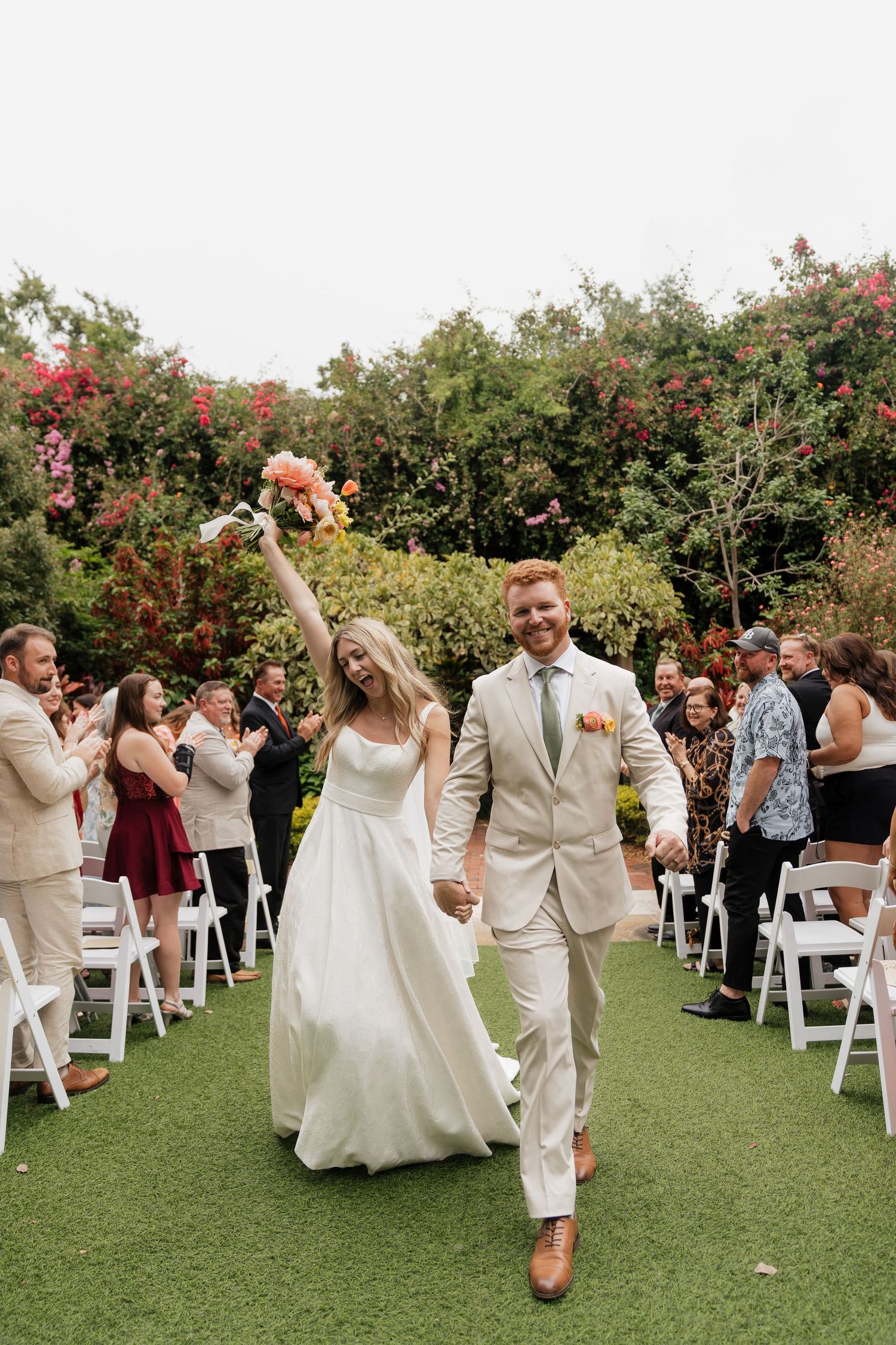 Wedding party celebrates joyfully with raised hands while walking down the aisle on a green lawn after outdoor ceremony.