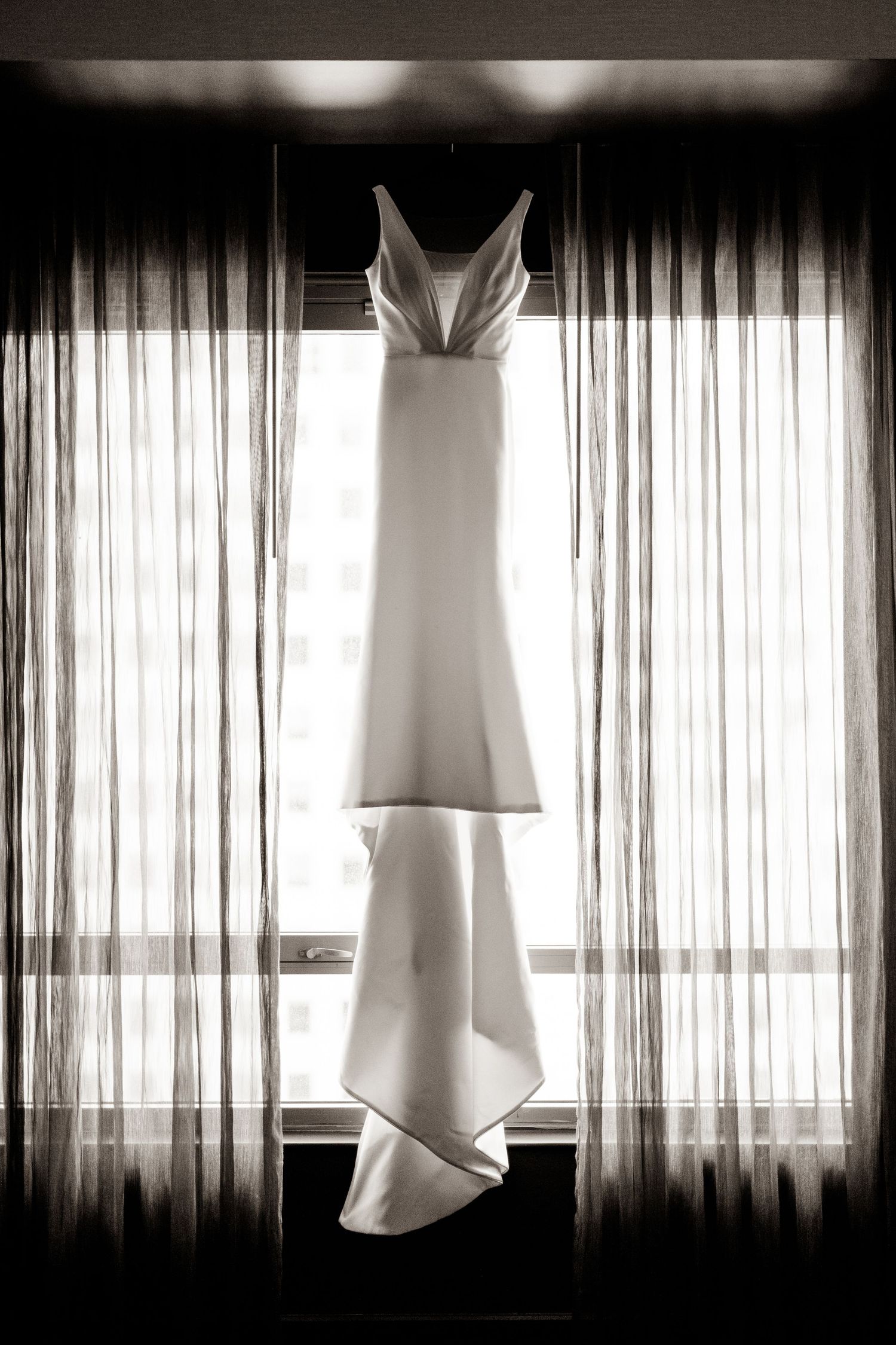 Elegant white wedding dress hanging in silhouette against sheer curtains in dramatic lighting.