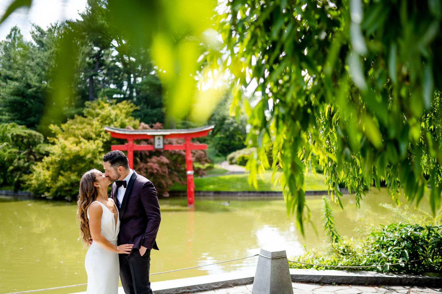 Wedding couple poses by Japanese garden pond with red torii gate in background.