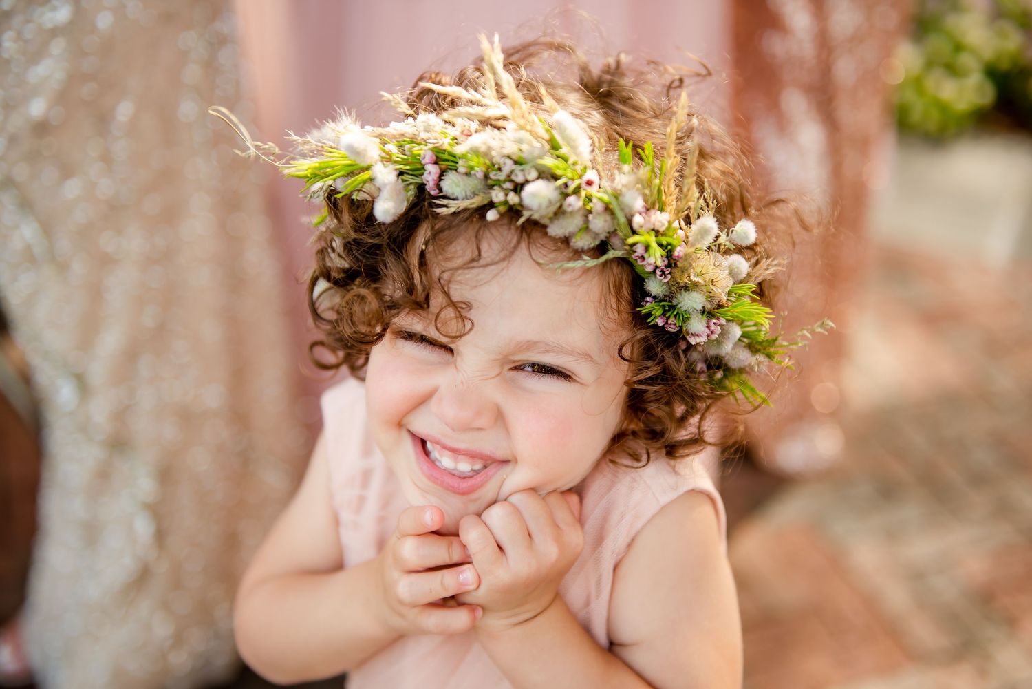 Young child wearing delicate floral crown smiles with hands on cheeks.