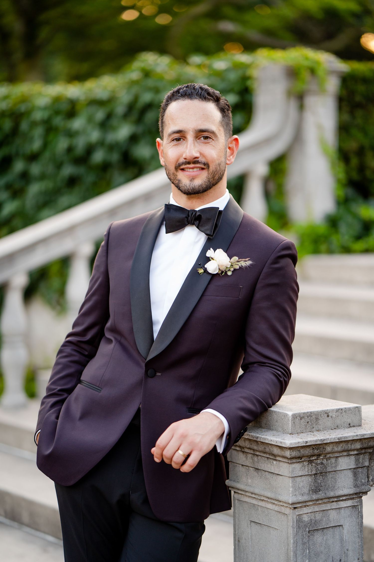 A groom in a burgundy tuxedo poses against a stone railing outdoors.